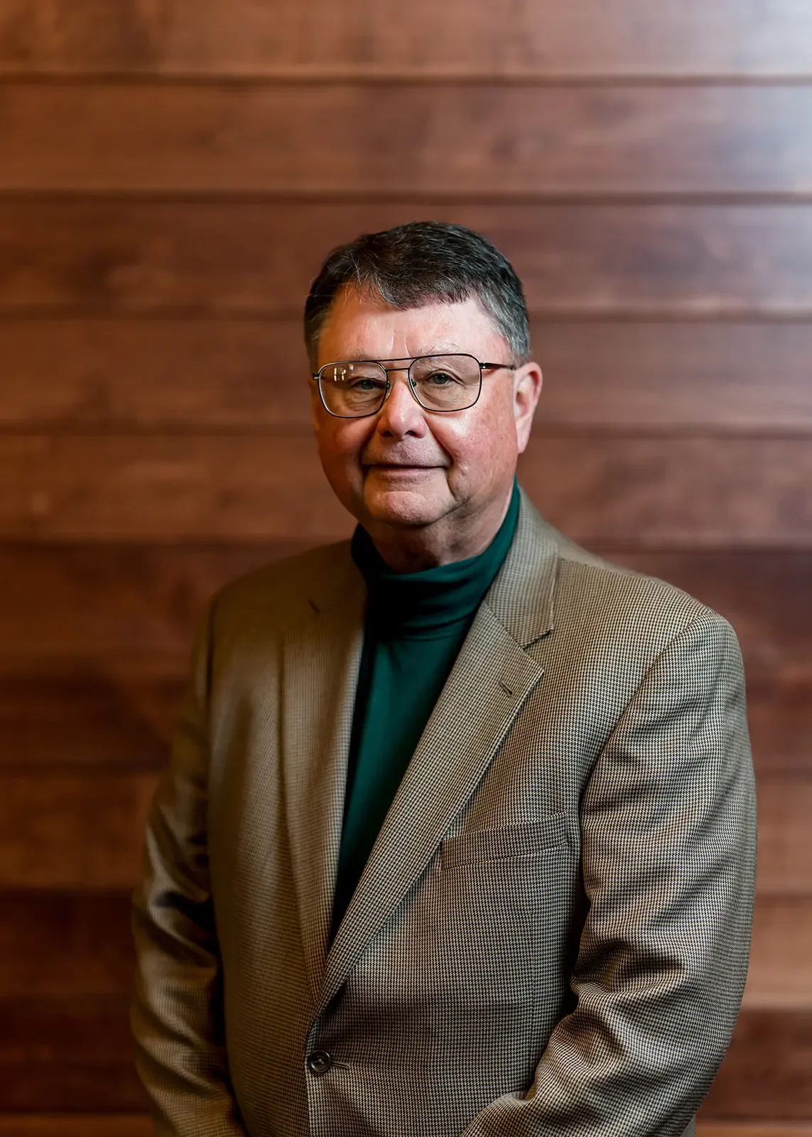 A middle-aged man with glasses wearing a brown blazer over a green turtleneck, standing in front of a wooden wall.