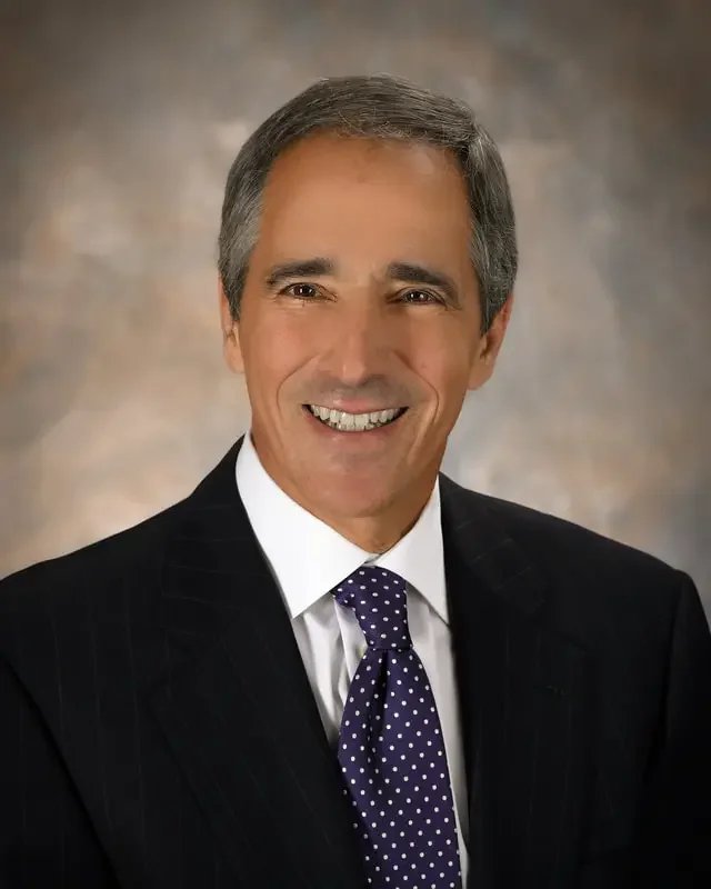 A middle-aged man with gray hair, wearing a dark pinstripe suit, white shirt, and purple polka dot tie, smiling against a brown and beige blurred background.