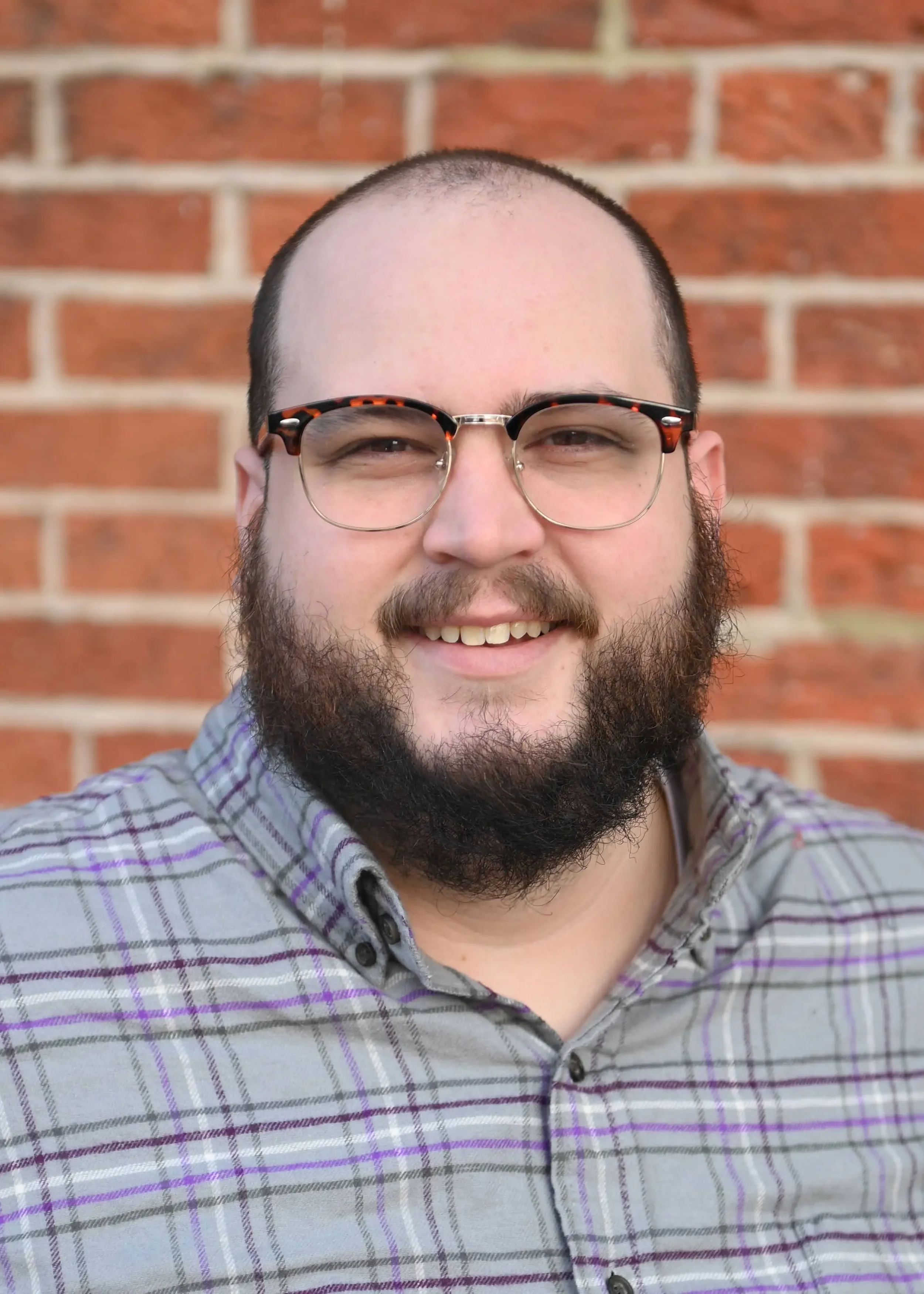 A smiling man with glasses, a beard, and short hair outdoors in front of a brick wall.