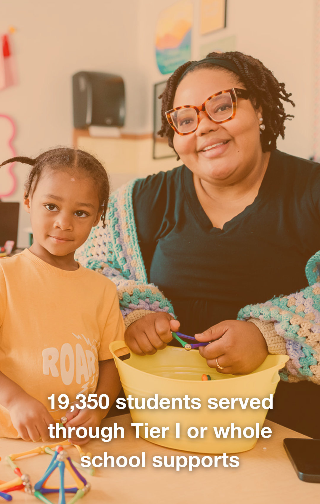 A woman and a young girl smiling at the camera, seated at a table with school supplies. The woman wears glasses and a colorful sweater, while the girl wears a yellow shirt and has dreadlocks. Text overlay reads, '16,083 students served through Tier 1 or whole school supports.'