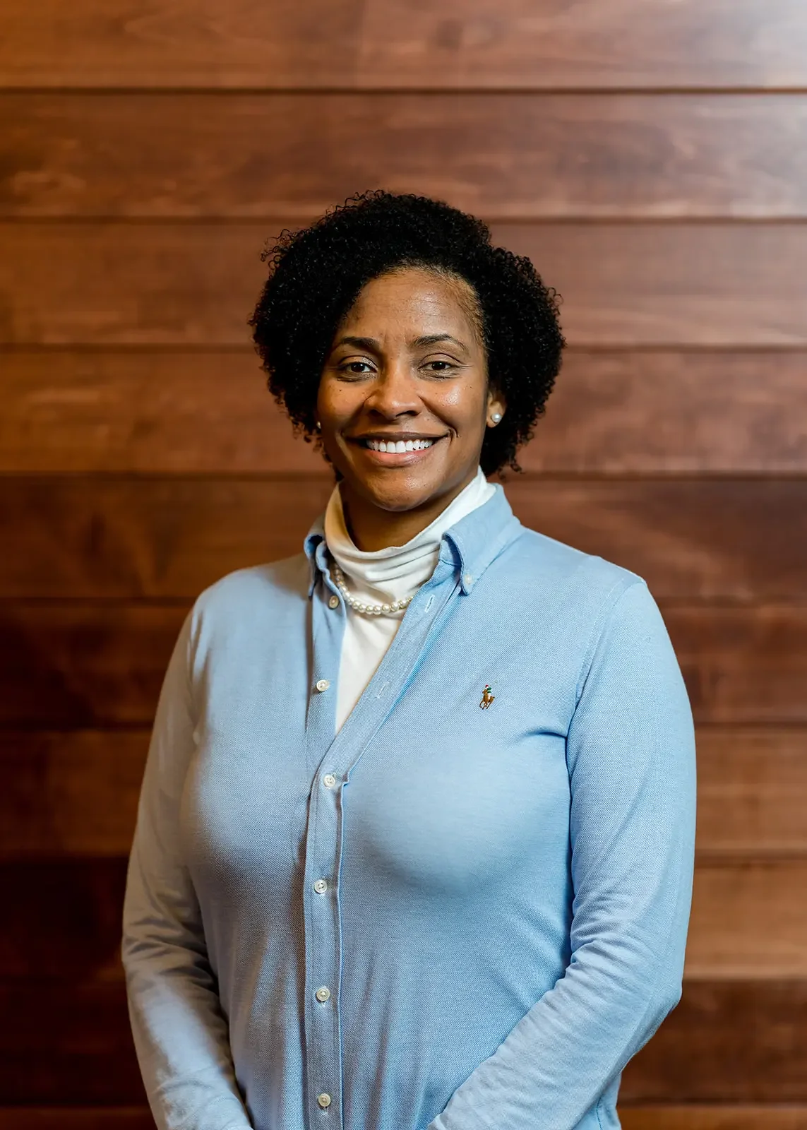 A woman with curly black hair smiling, wearing a light blue button-up shirt over a white turtleneck, standing in front of a wooden wall.