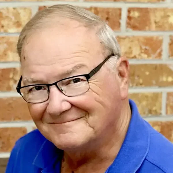 A smiling middle-aged man with glasses and short gray hair, wearing a blue shirt, standing in front of a brick wall.