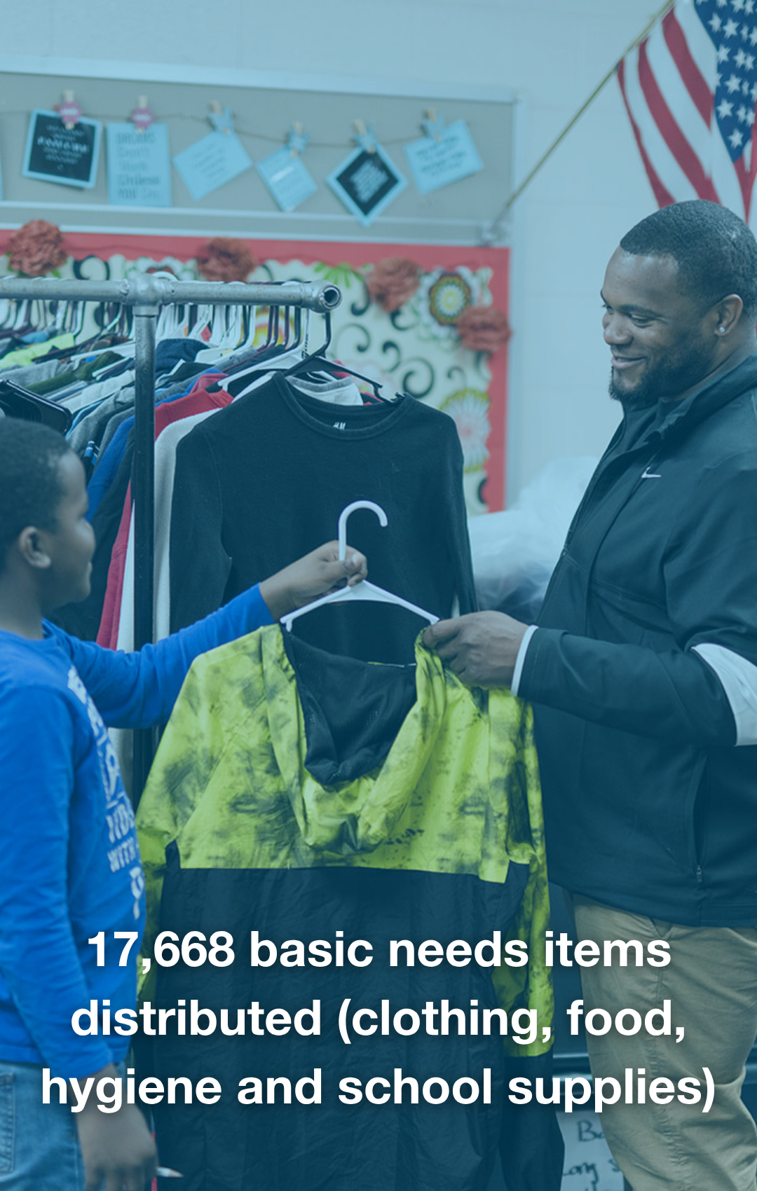 A man and a boy shopping for clothing at a donation event, with a rack of clothes in the background and an American flag in the corner.