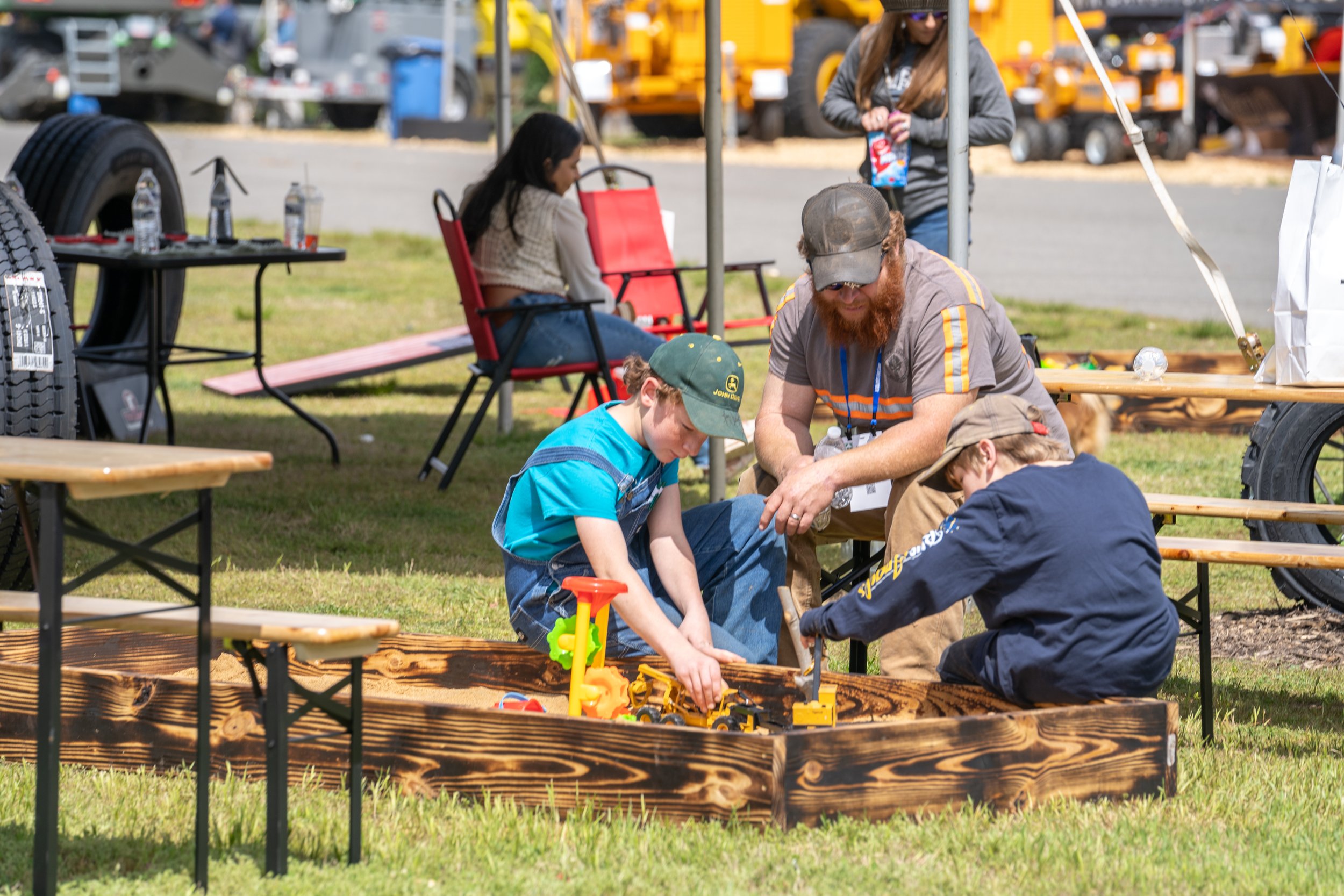 Three children and two adults playing with toy construction vehicles in a wooden sandbox at an outdoor event with tents, chairs, and various equipment in the background.