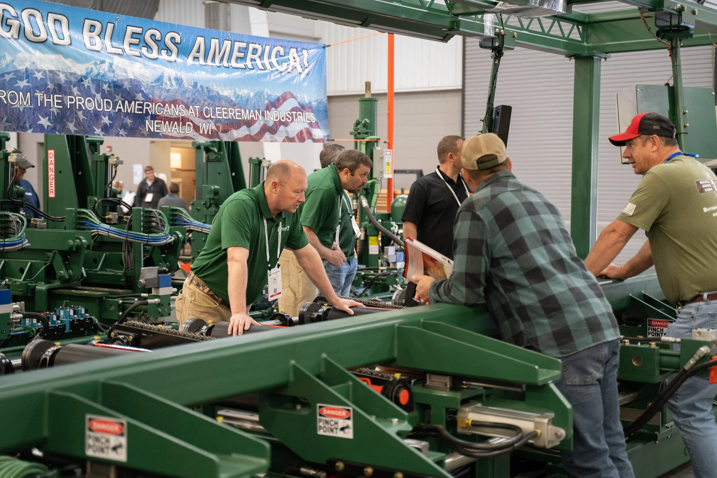 Group of men examining green machinery at a trade show booth with a patriotic banner that reads 'God Bless America'.