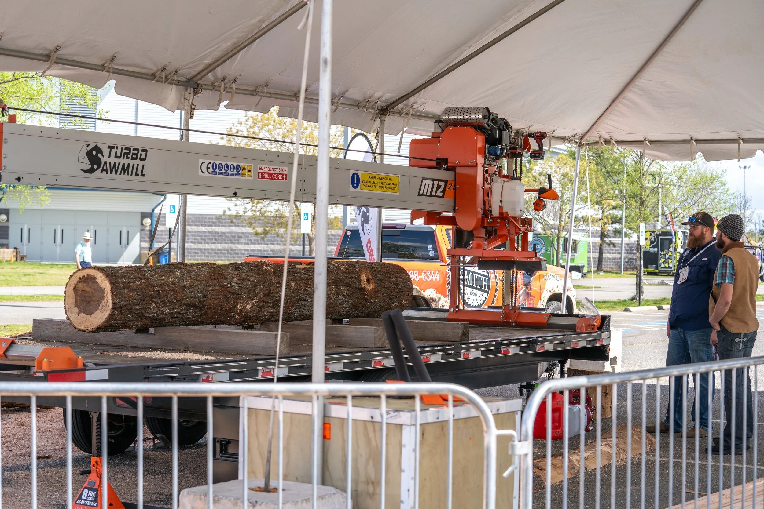 A large log is placed on a conveyor belt under a mechanical saw in a tented outdoor setting, with two men observing the equipment.