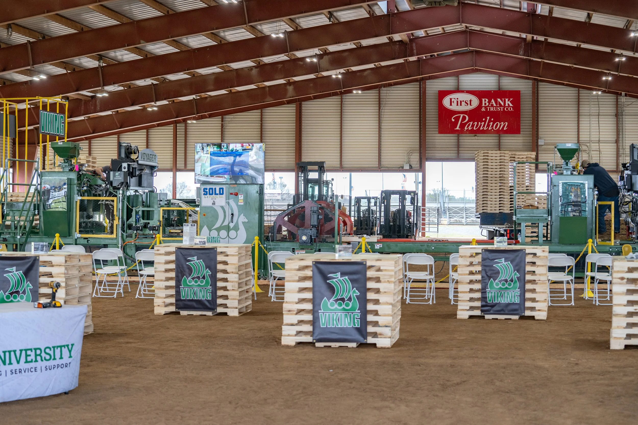 An indoor venue with industrial-style architecture, featuring a central display of vintage printing presses, wooden pallets, and promotional banners for Viking and a university, under a large metal arched roof with a sign reading "First Bank Pavilion