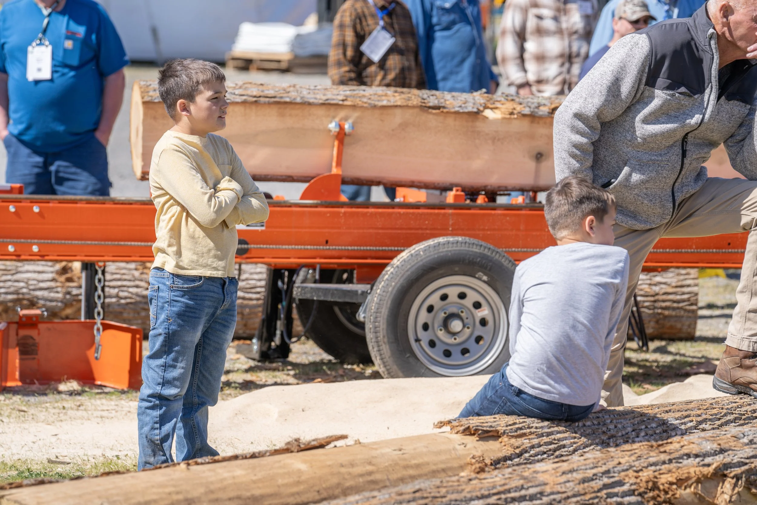 Children and adults gathered outdoors near logs and sawmill equipment, with a boy standing with crossed arms and others sitting or leaning around a large log on a trailer.