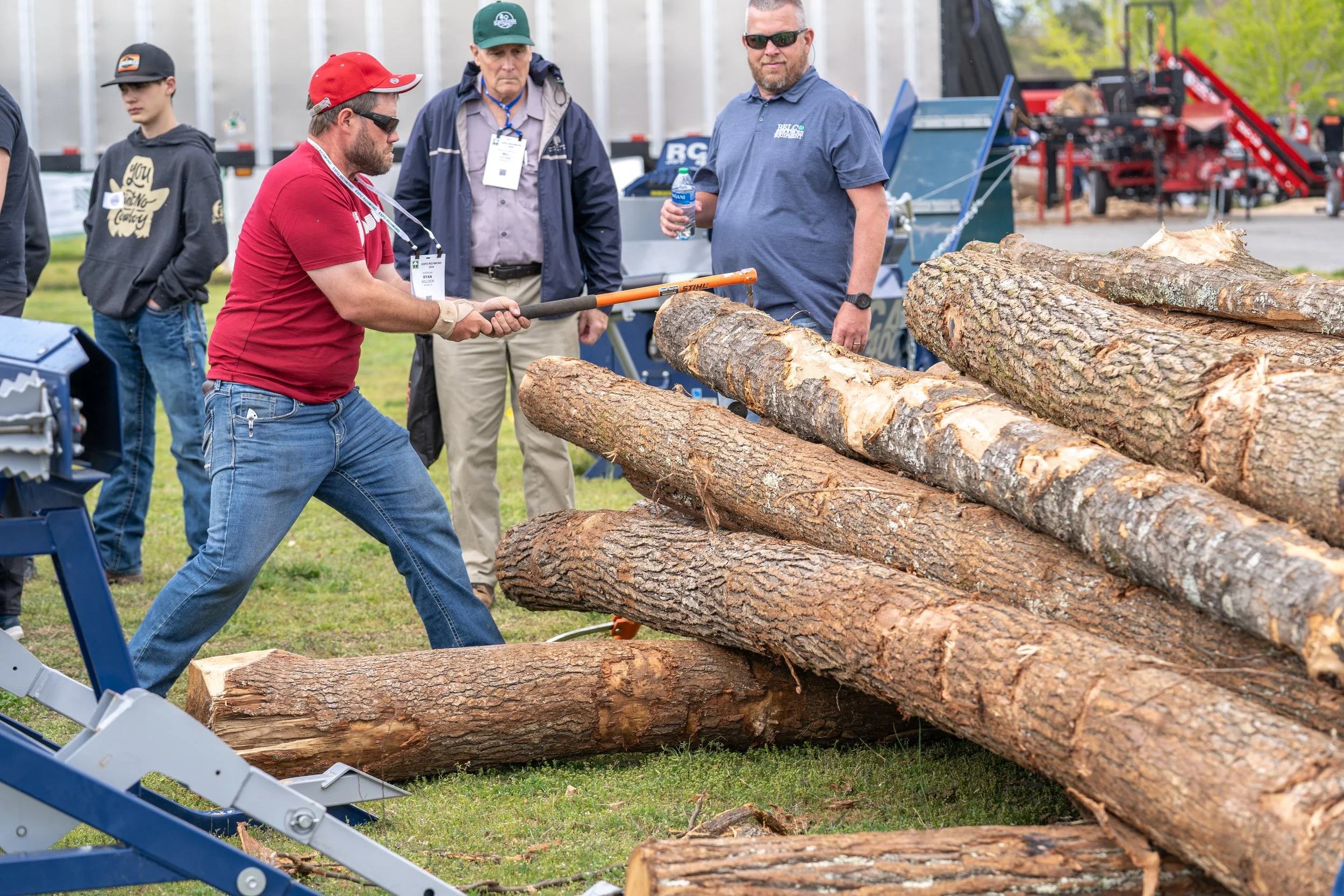 Men cutting logs with saws at an outdoor event, with onlookers observing.