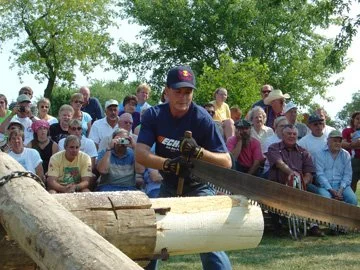 A man in a navy blue shirt and baseball cap demonstrating chainsaw carving to a seated crowd outdoors.