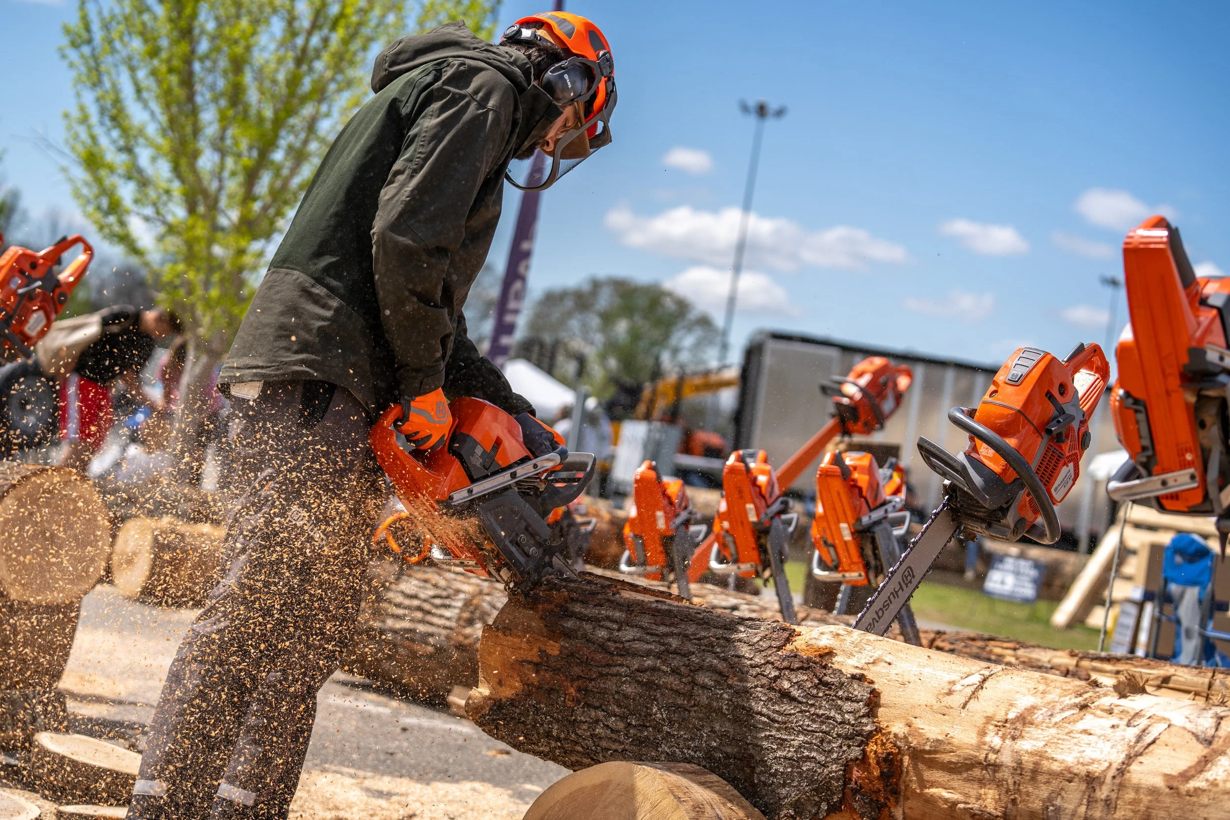 A person wearing safety gear and orange gloves is using a chainsaw to cut a log of wood at an outdoor event, with multiple chainsaws lined up nearby.