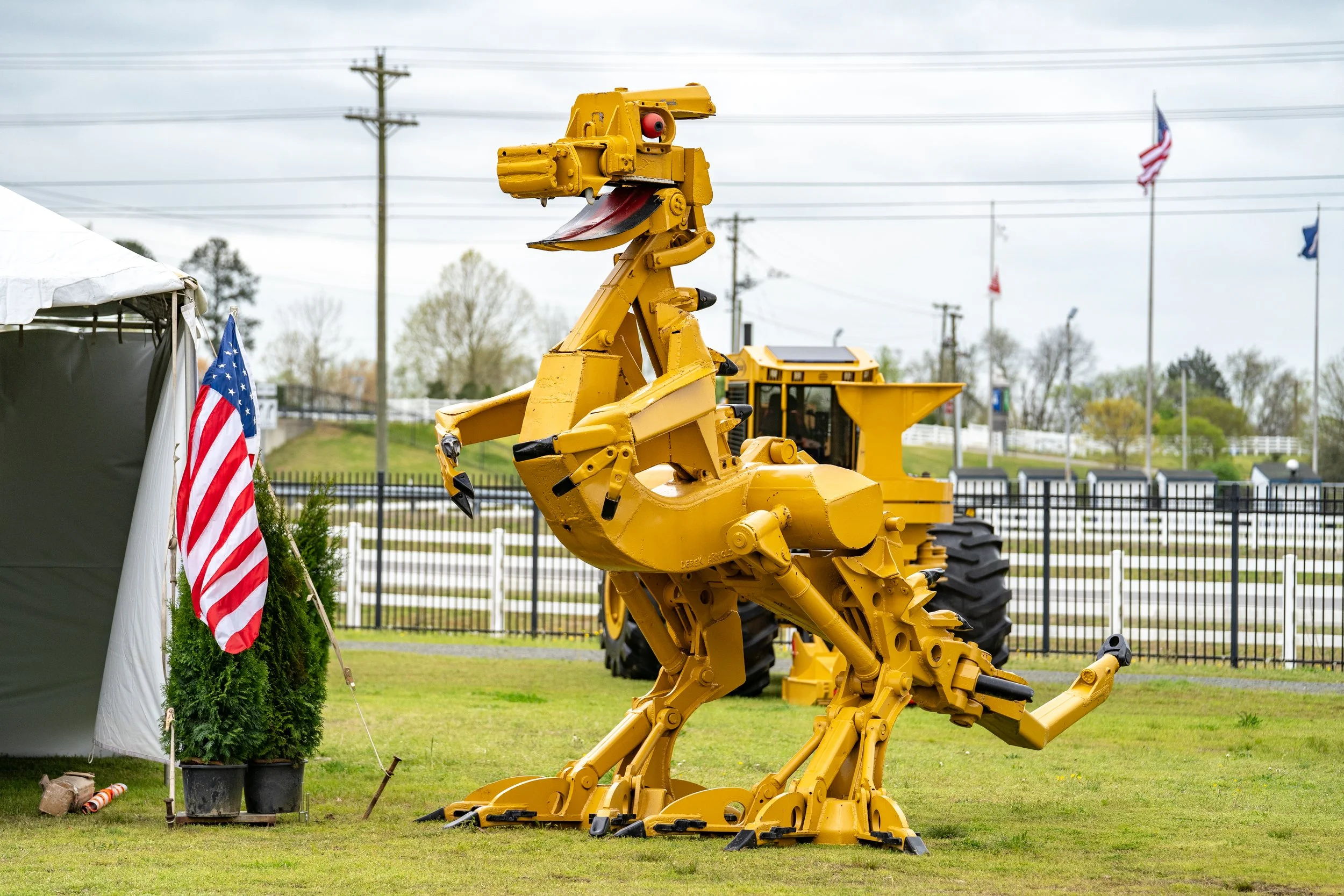 A mechanical dinosaur sculpture painted yellow, set outdoors on grass with a tent and American flags nearby, and a yellow construction vehicle in the background.