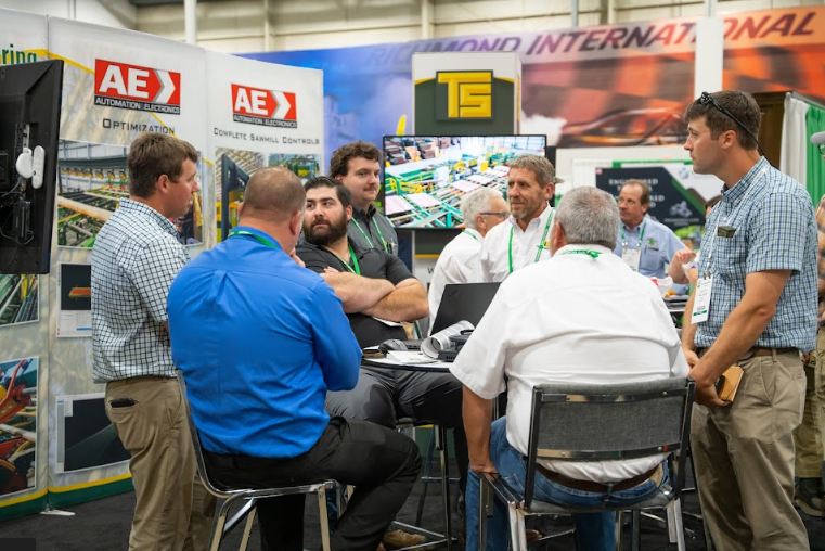 Group of men gathered around a table at a trade show booth, discussing among displays with signage that reads 'AE' and 'T5'.
