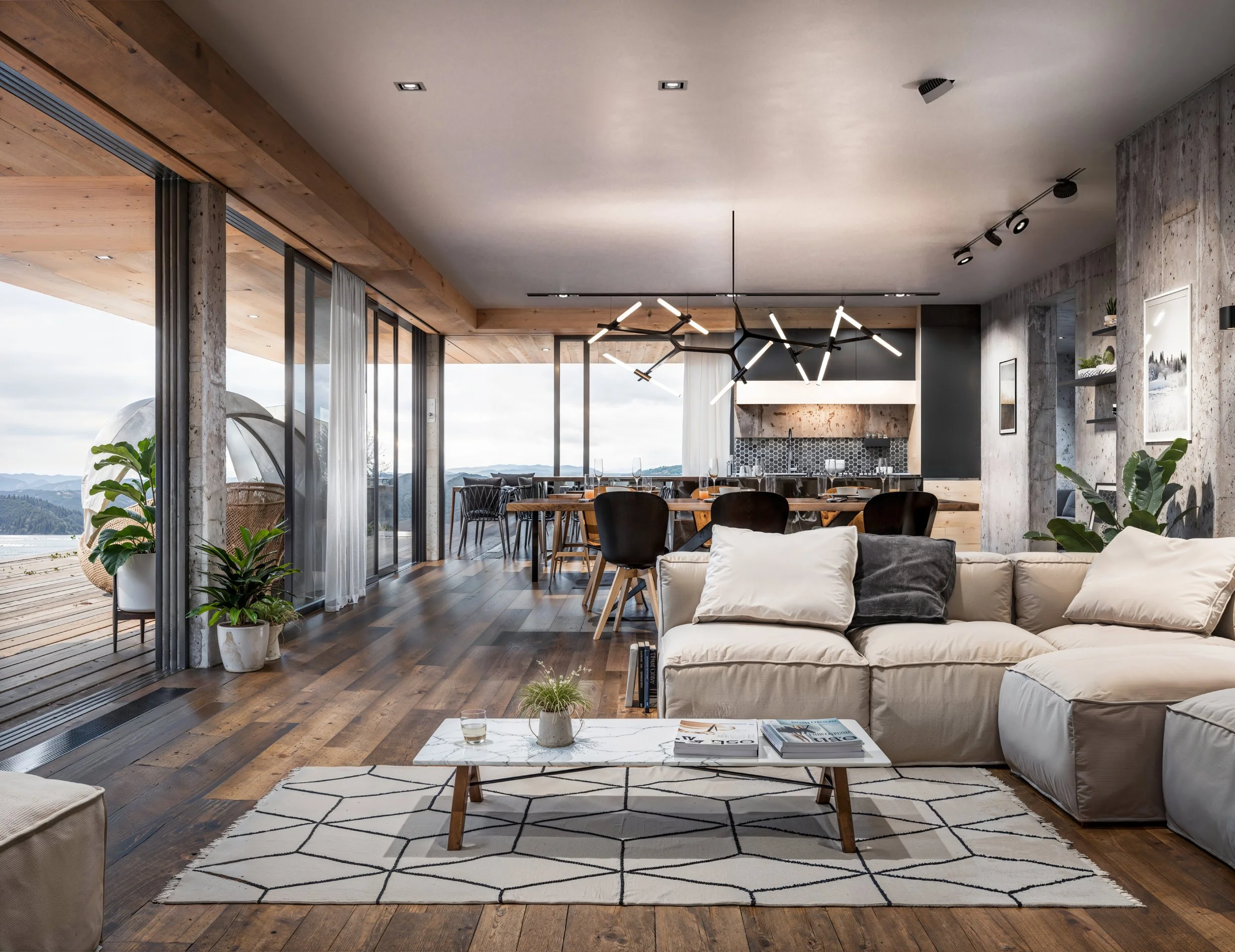 Modern living room with large windows overlooking mountains, featuring a beige sectional sofa, a marble coffee table, wooden dining table with black chairs, contemporary chandelier, plants, and a kitchen with patterned backsplash in the background.
