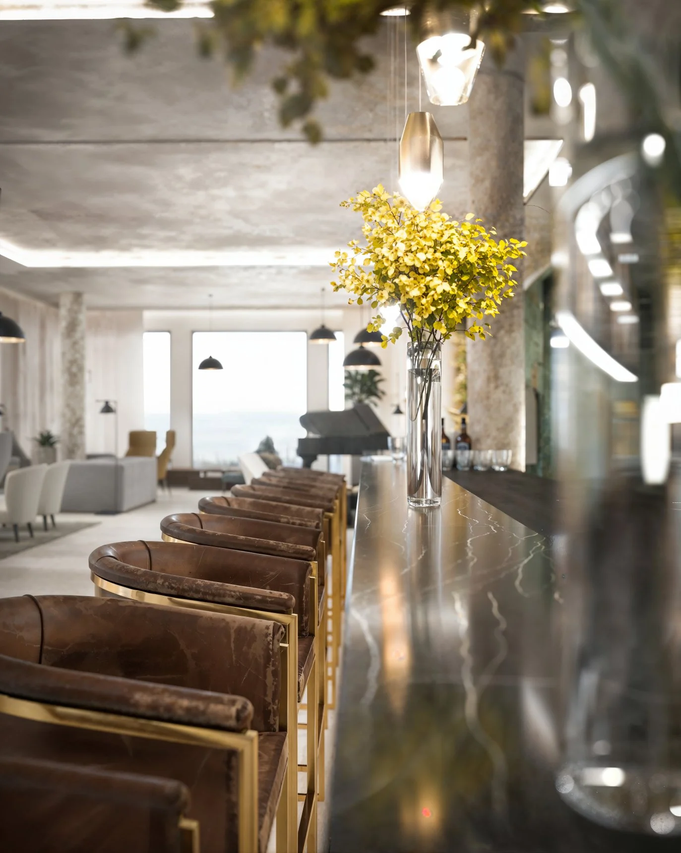 Interior of a modern lounge or bar area featuring a marble countertop with a glass vase holding yellow flowers, leather chairs with gold frames, and pendant lights hanging from the ceiling, with large windows in the background.