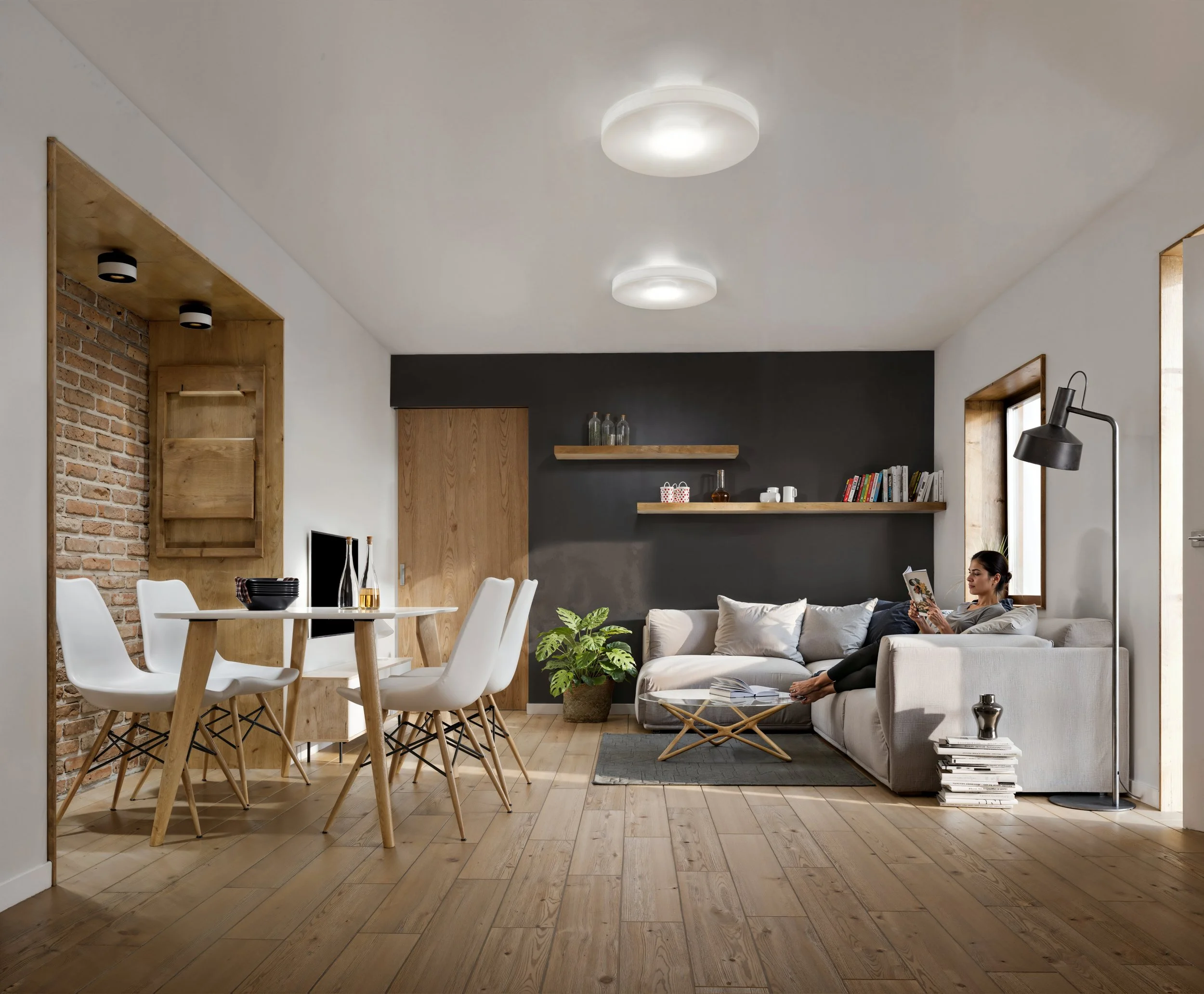 Open-concept living and dining room with white walls, wooden flooring, and modern furniture. A woman sits on a light-colored sectional sofa reading a magazine near a floor lamp. The dining area has a round wooden table with white chairs. There's a bl