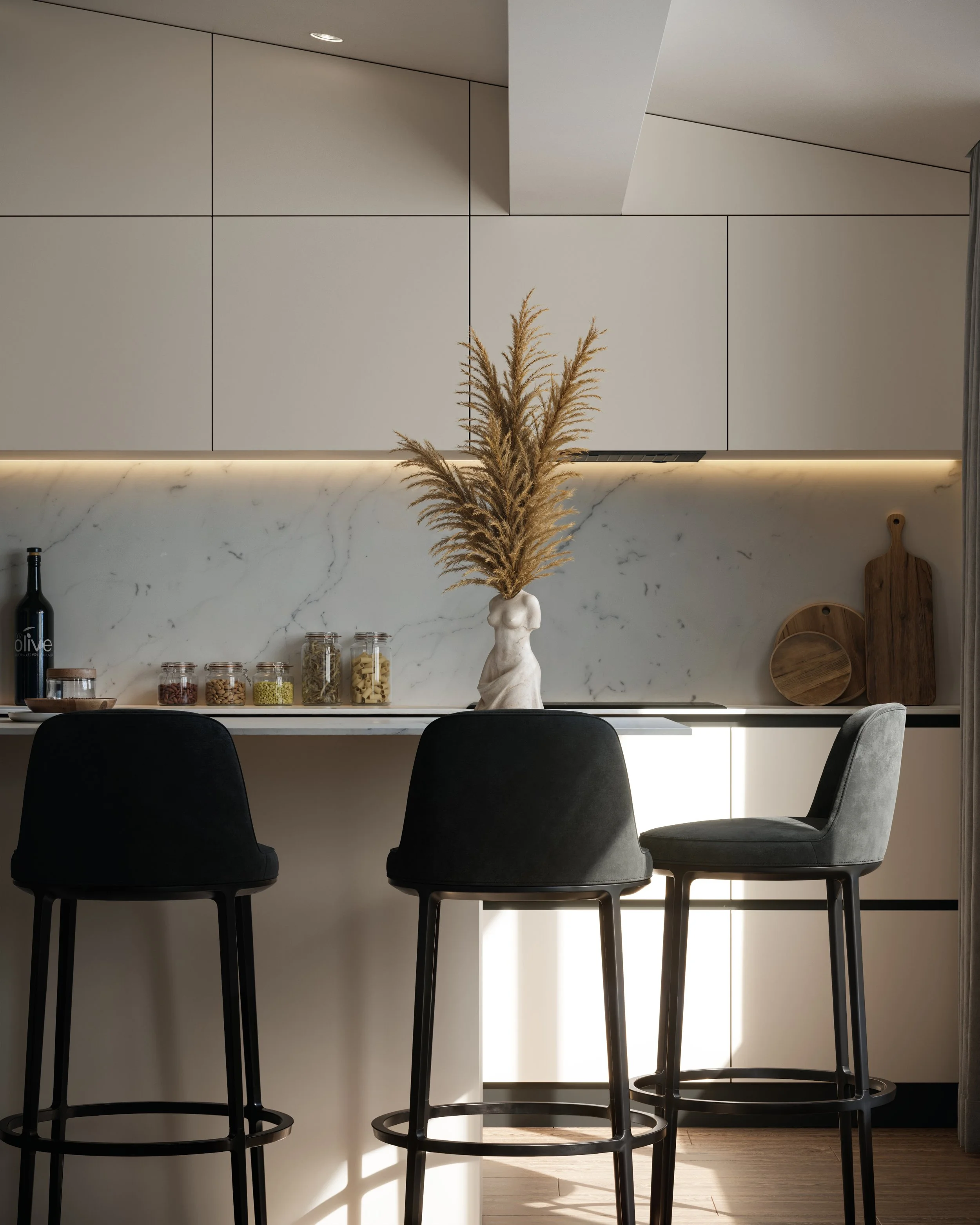 A modern kitchen with beige cabinets, marble backsplash, and three black bar stools at a white countertop. A vase with dried pampas grass is on the counter, along with jars of food, and wooden cutting boards.