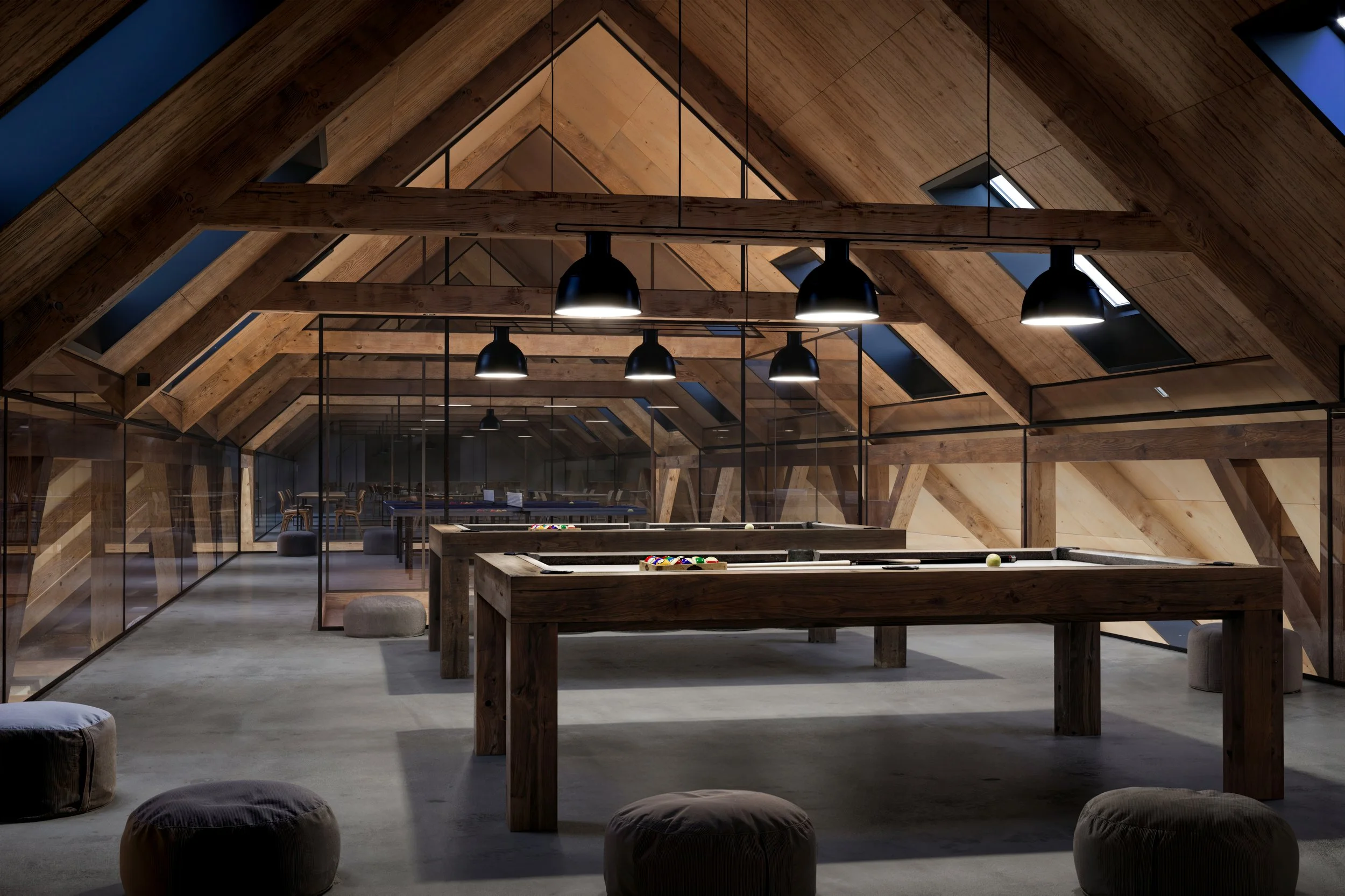 An attic game room with wooden A-frame ceiling and modern decor, featuring pool tables, hanging black pendant lights, and pouf seating on a concrete floor.