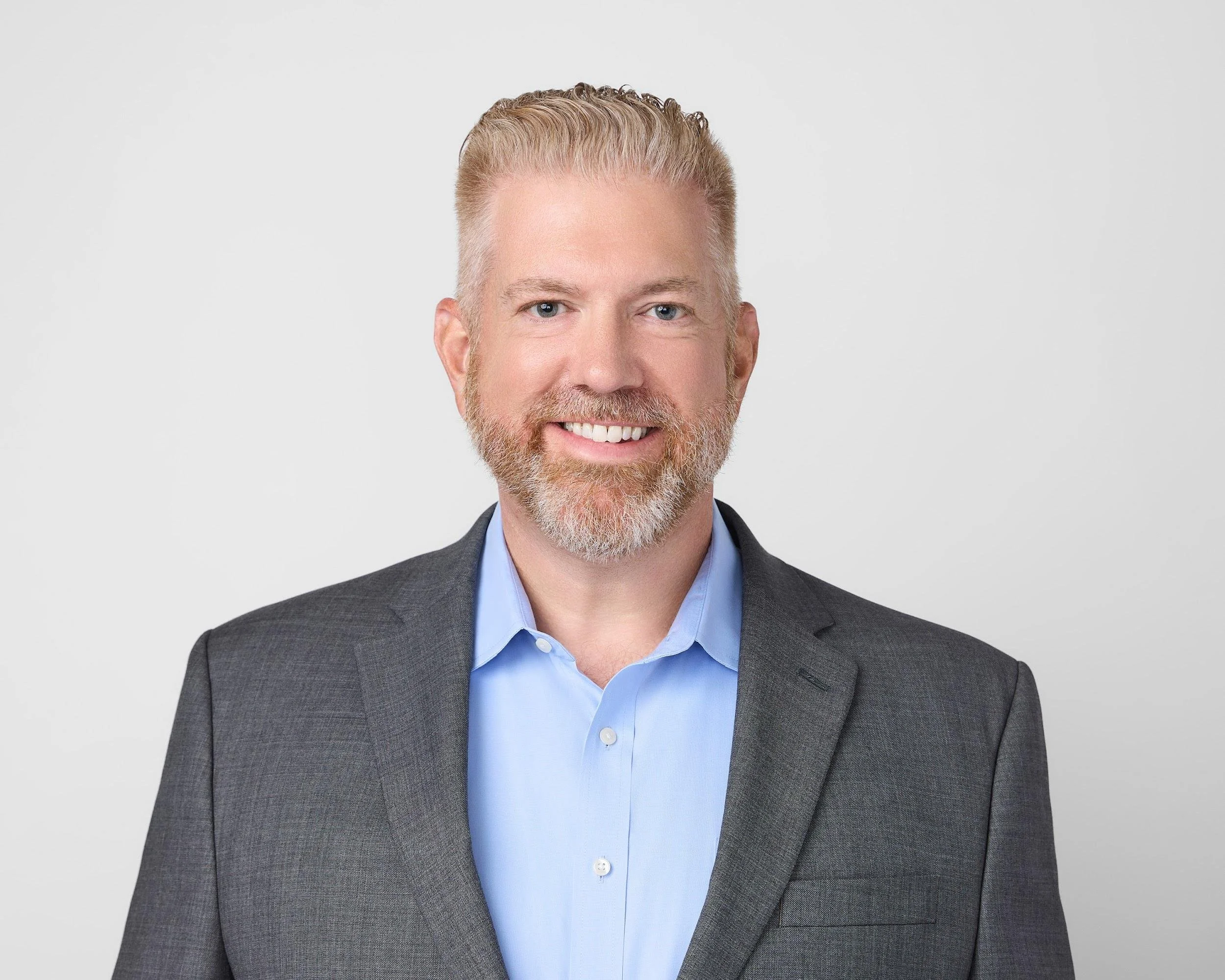 A smiling man in a gray suit jacket and light blue dress shirt against a plain white background.