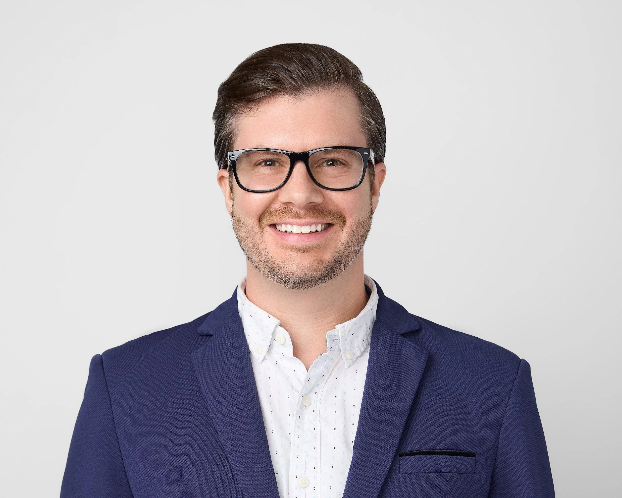 A smiling man with brown hair, glasses, and a beard, wearing a navy blazer over a white shirt with small pattern, against a plain light background.