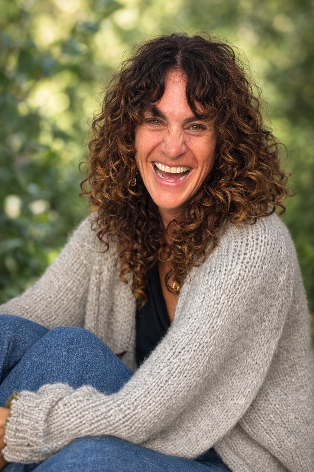 Meryl Fields sitting outdoors under green trees, smiling and wearing a gray knit sweater and blue jeans.