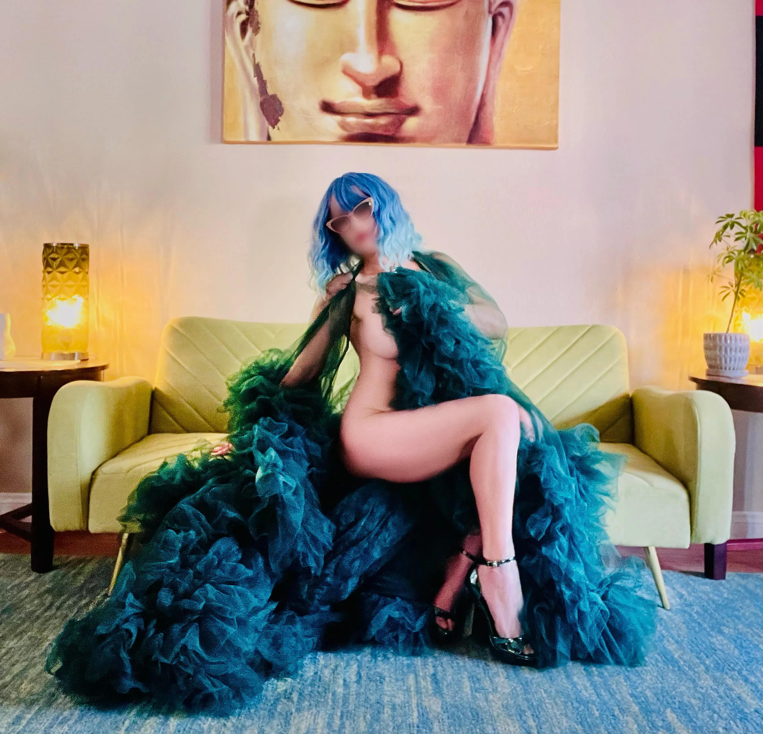 Vegas woman with blue hair and sunglasses sits on a couch wearing a large dark green ruffled robe, revealing her breast and legs. The background features a Buddha painting on the wall, and there are two side tables with lamps and plants.
