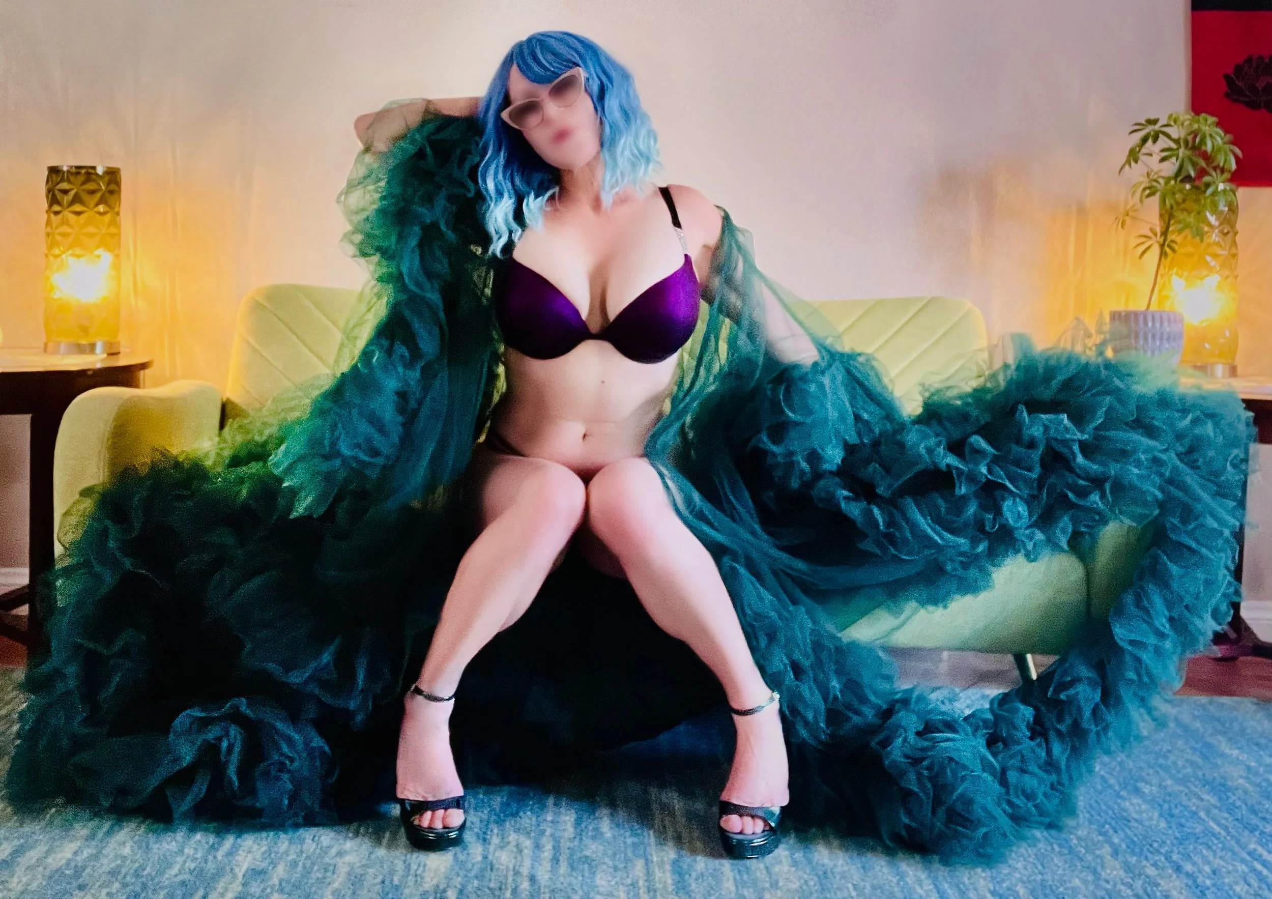 Las Vegas Nuru expert blue hair, wearing sunglasses, a purple bra, high heels, and a voluminous green dress with ruffles, sitting on a green couch in a warmly lit room with lamps and indoor plants.