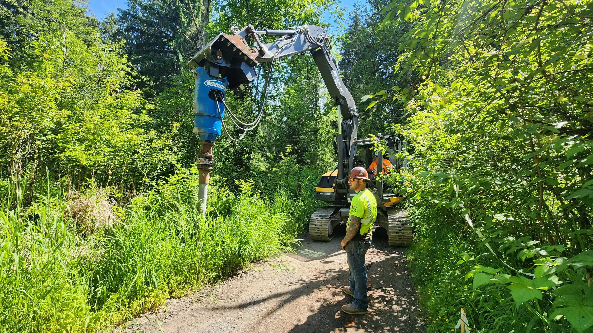 A man in a neon shirt stands next to a large machine with a drill that's being operated by another man.