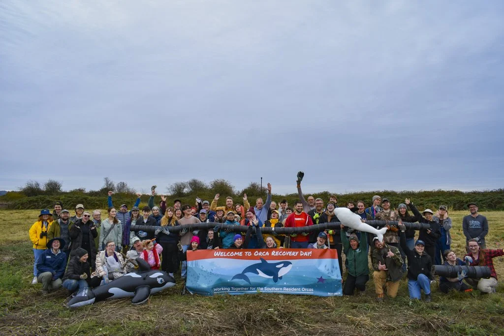 Group of smiling people holding a banner that says "welcome to Orca Recovery Day!" Some are holding plant pots and orca toys