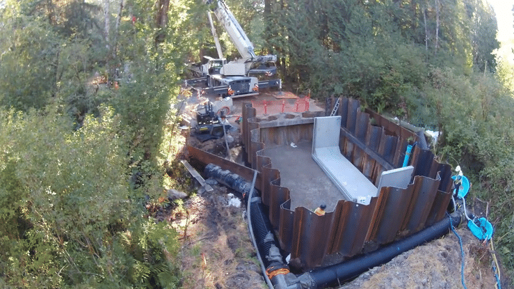 A repeating timelapse of a crane lowering concrete shaped like a bracket into the ground. People with hardhats and safety gear move throughout the frame.
