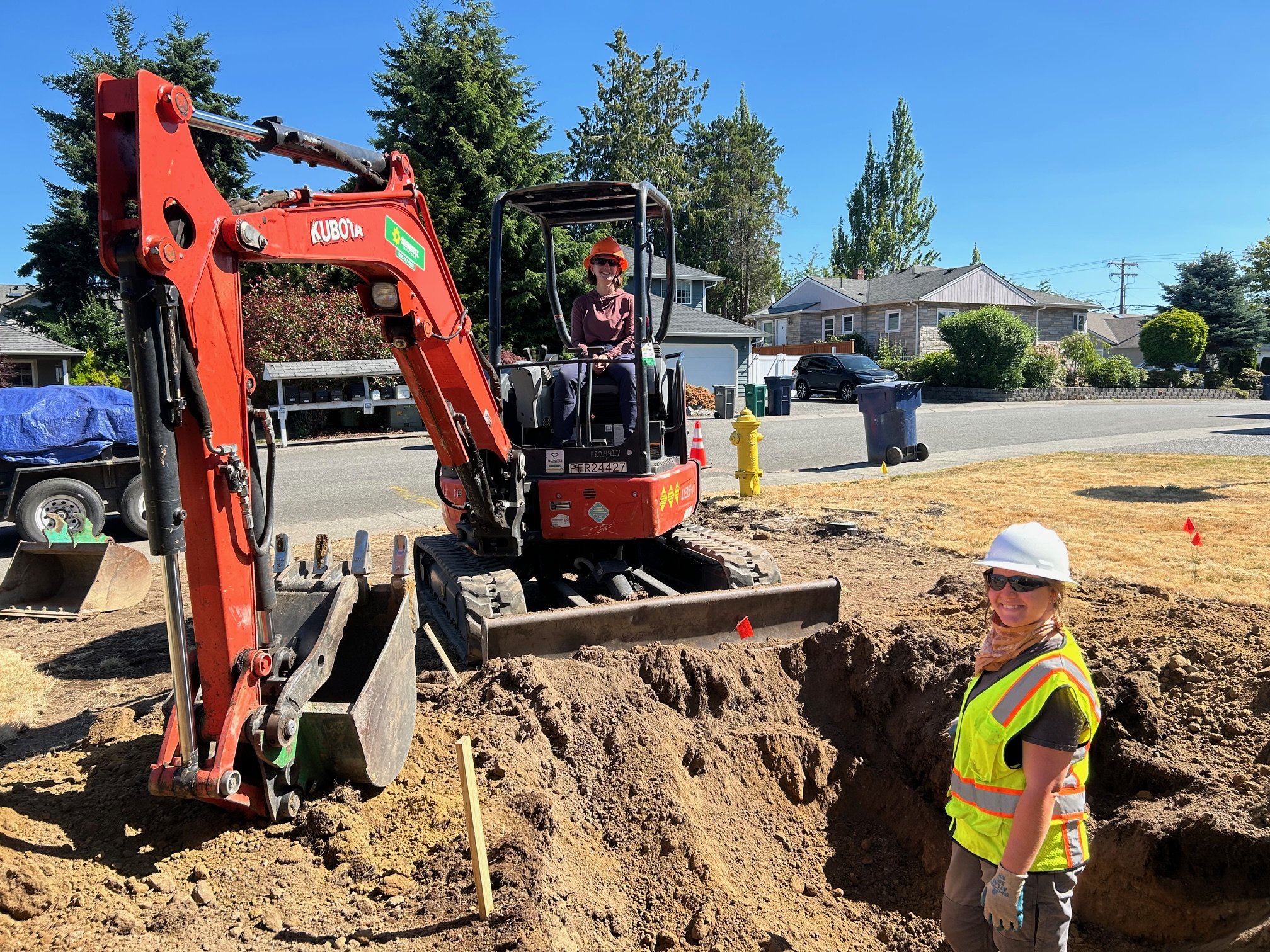 Two people wearing reflective vests and hardhats smile while digging with an excavator