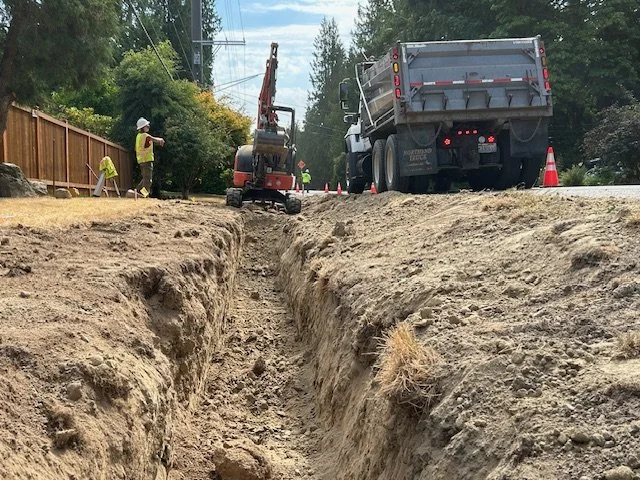 A deep trench alongside a road with an excavator and large truck in the background.