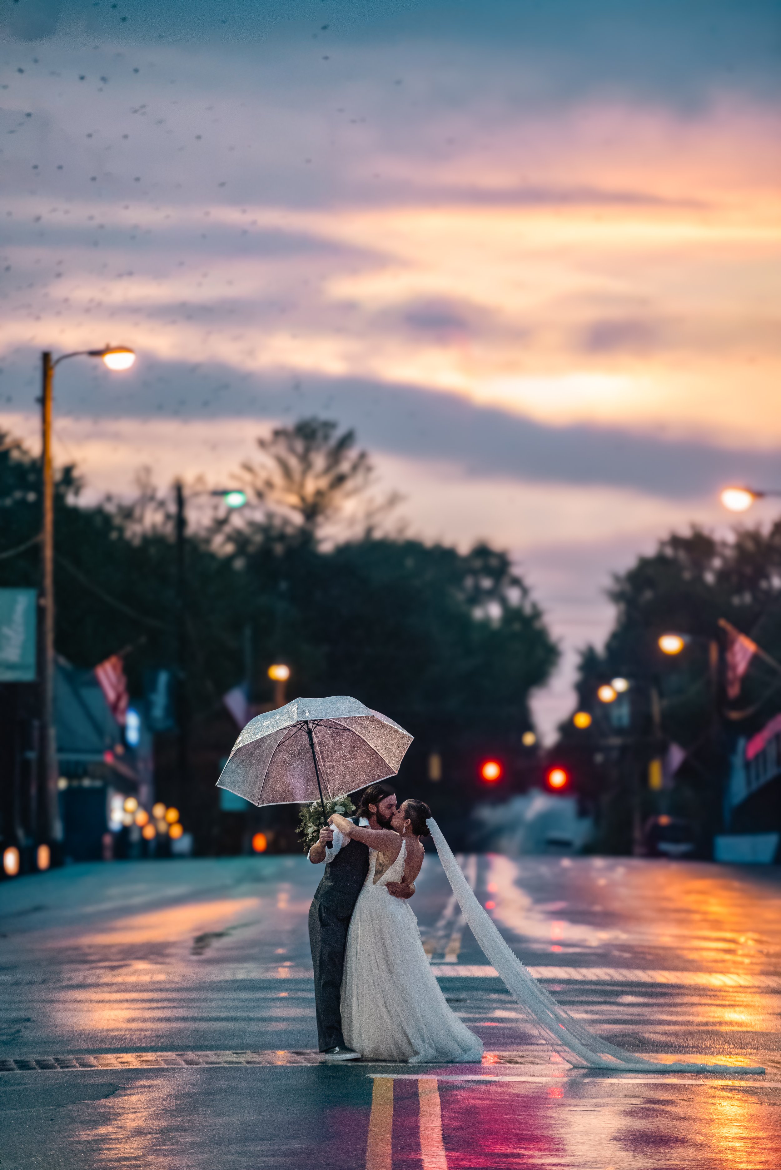 A bride and groom sharing a kiss under an umbrella on a rainy street during sunset.