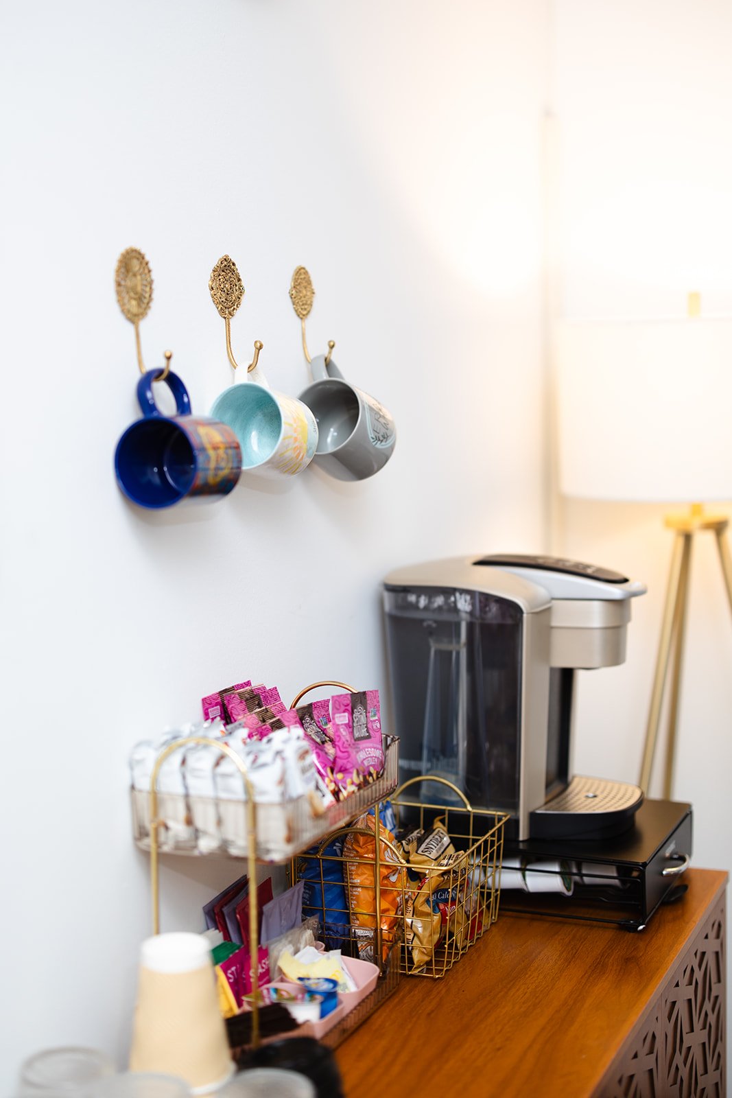 Coffee mugs hanging on gold hooks on a white wall next to a coffee station with a Keurig machine and snack baskets on a wooden counter.