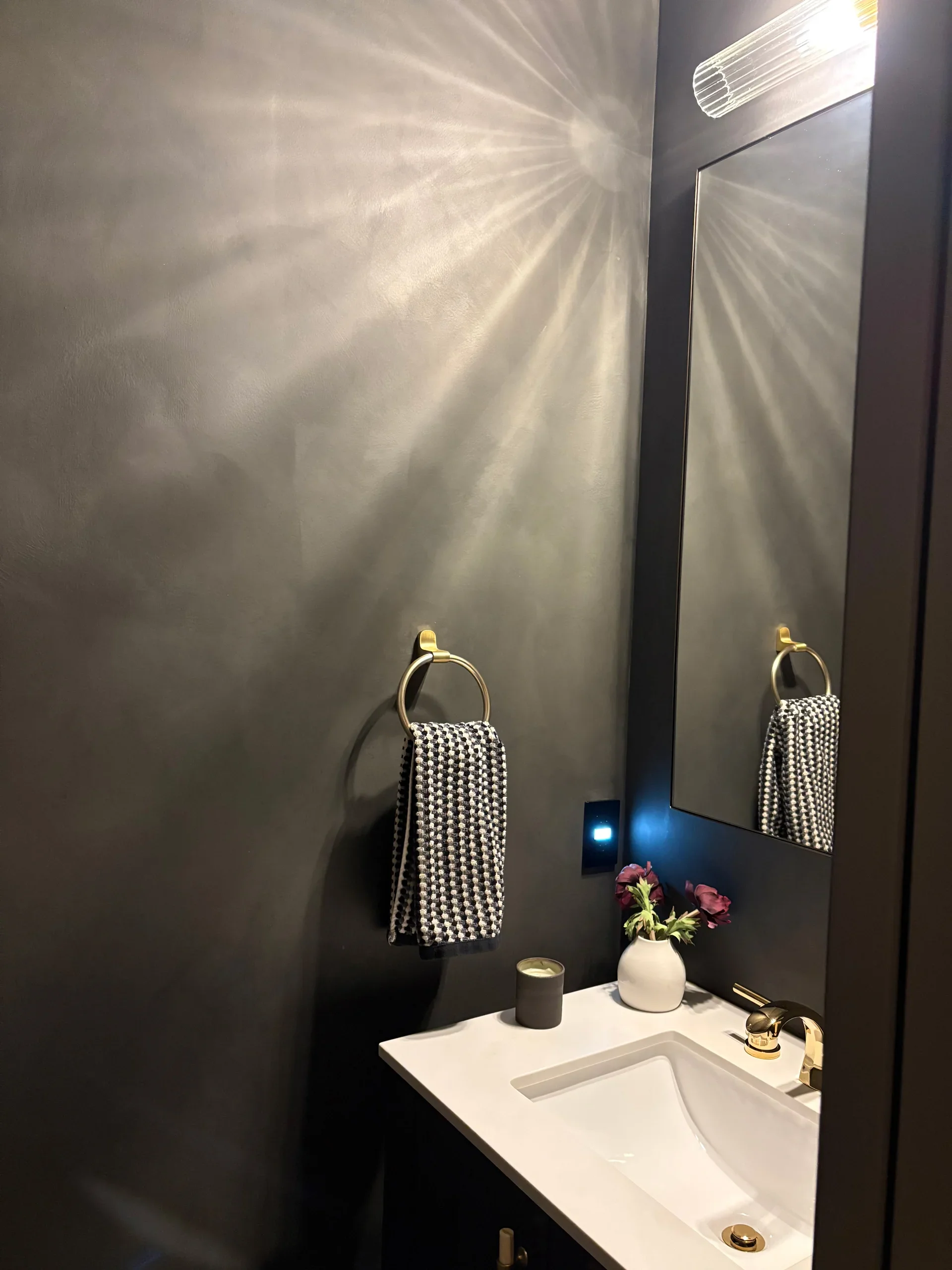 A modern bathroom with a black wall, gold towel ring holding a black and white patterned towel, a white countertop with a sink, a small candle, and a vase with purple flowers. A mirror reflects the bathroom light.