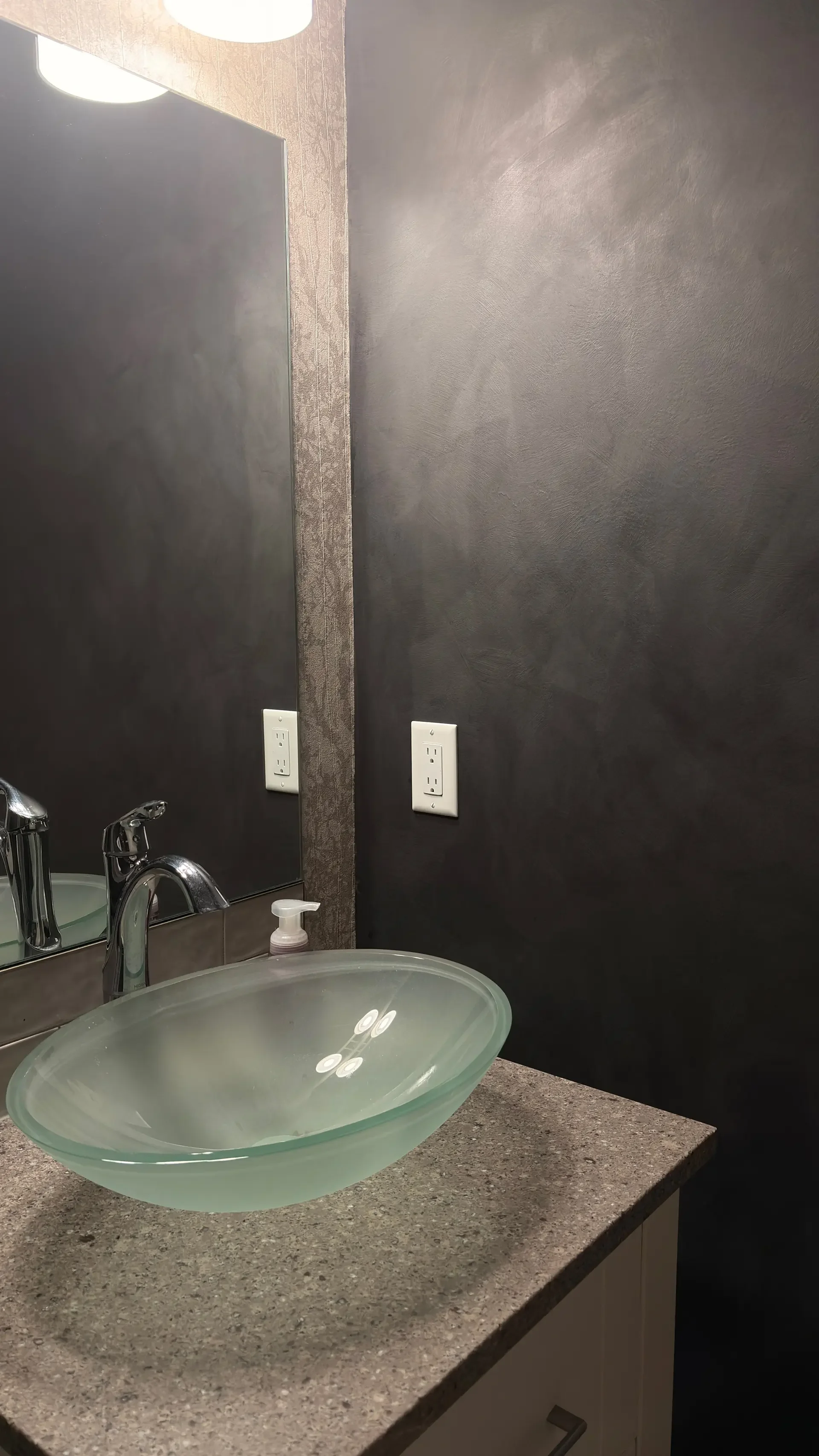 Bathroom sink with a frosted glass vessel bowl, soap dispenser, granite countertop, large mirror, dark textured wall, and electrical outlets.