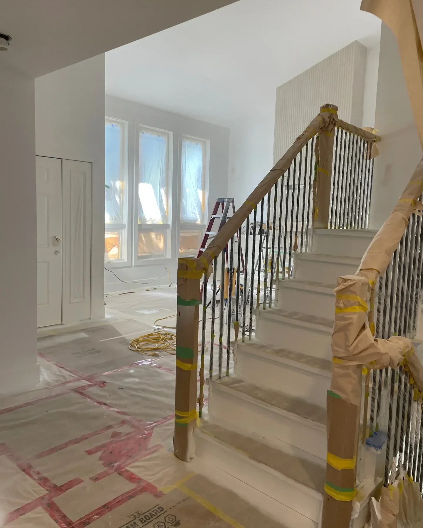 Interior of a house under construction or renovation, featuring a staircase with new handrails covered in brown paper and yellow tape, a ladder, and plastic sheeting on the floor to protect surfaces.