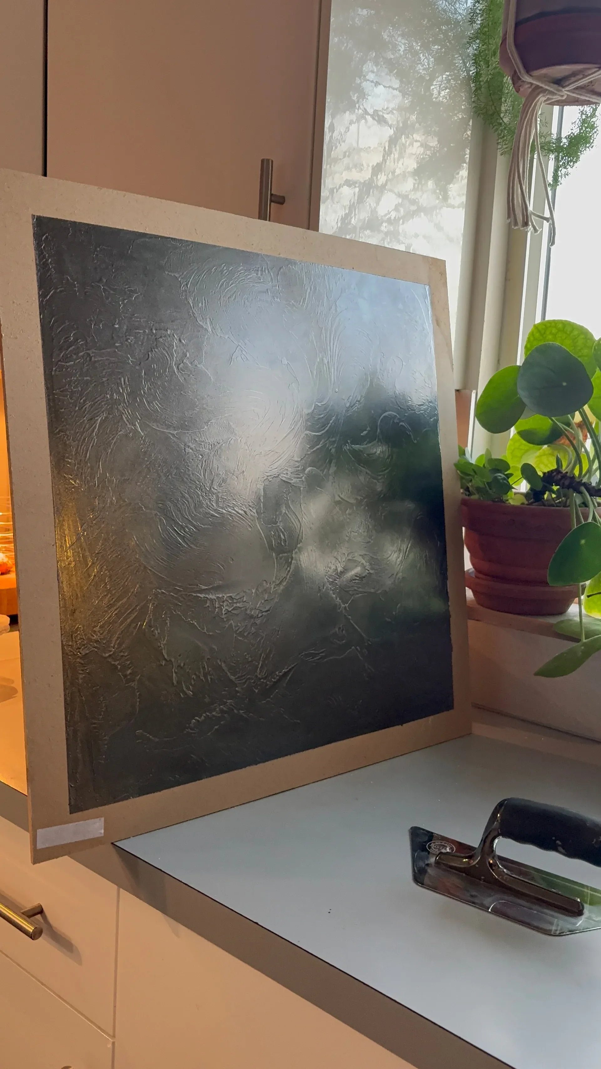 A metal sheet with engraved or textured abstract patterns, resting on a white countertop next to a black and silver iron. There are green plants in a reddish-brown pot and a window with a view of trees in the background.