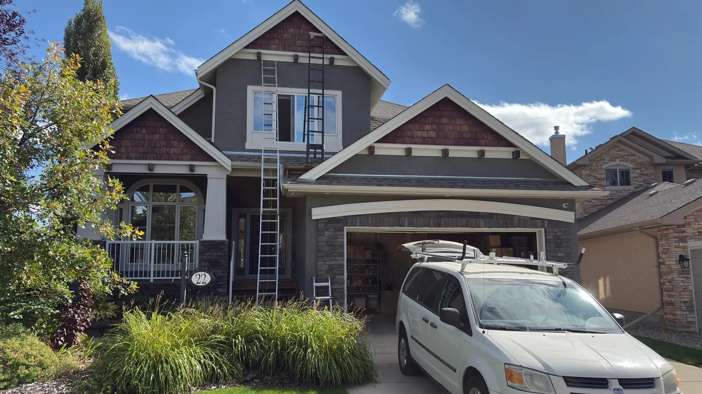 A two-story house under construction with ladders on the roof, a white van parked in the driveway, and lush green landscaping in front.