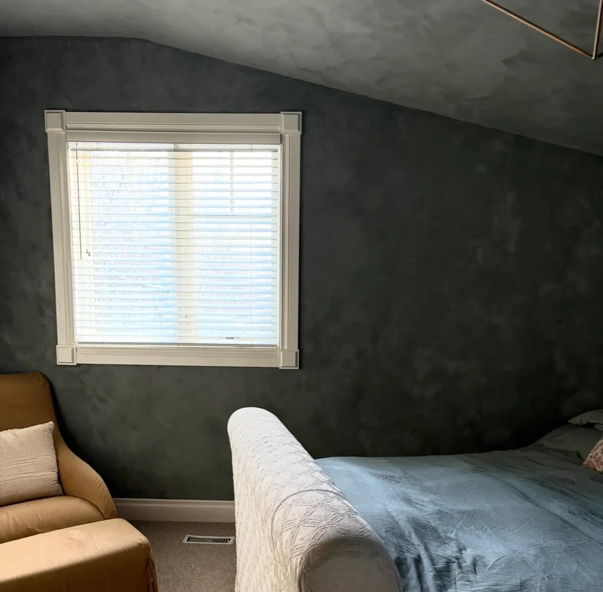 A cozy bedroom with a dark textured wall, a window with white blinds, a bed with a gray comforter, and a beige armchair with a pillow.