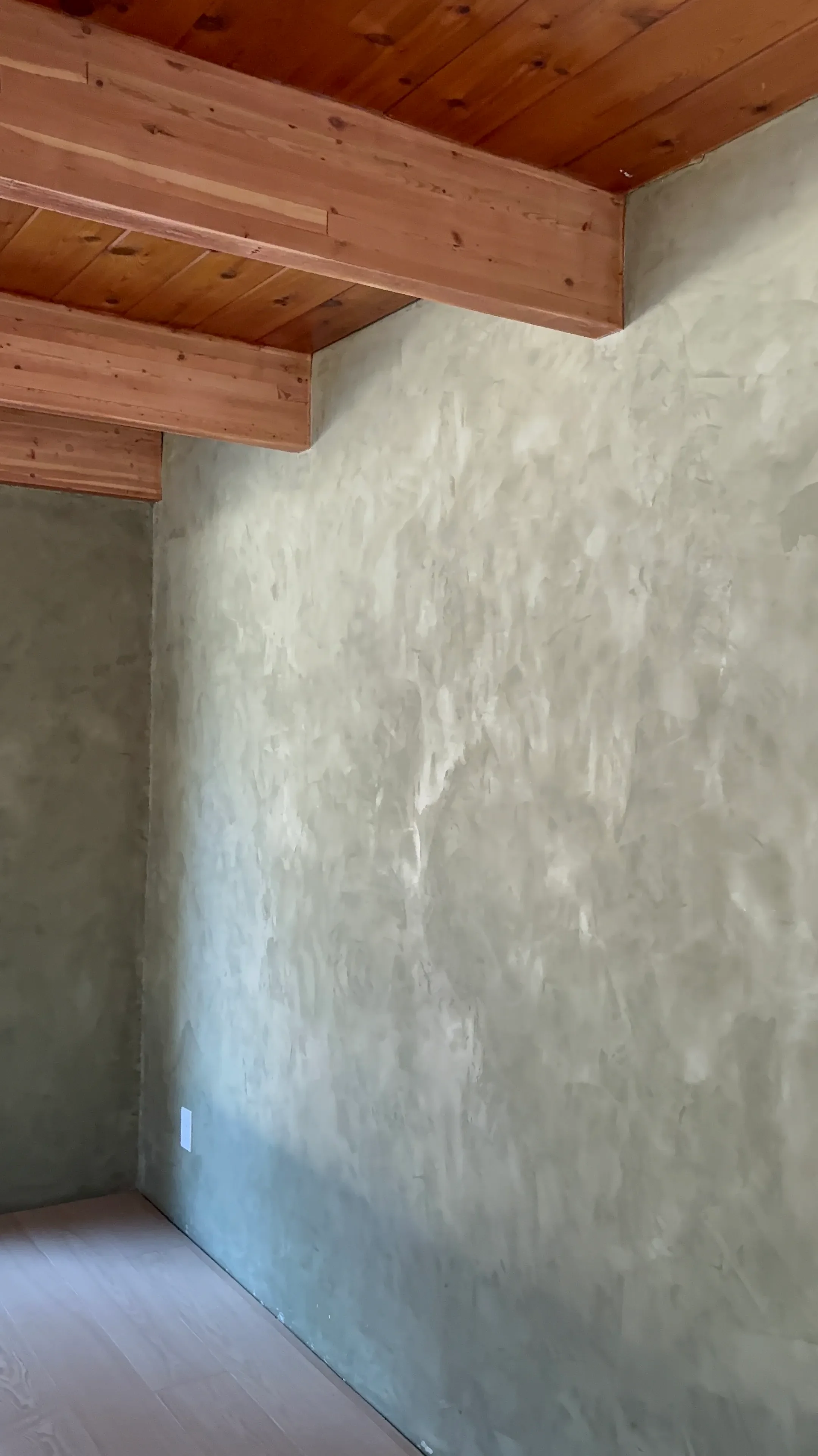 Interior corner of a room with a textured beige wall, wooden ceiling beams, and a light wooden floor.