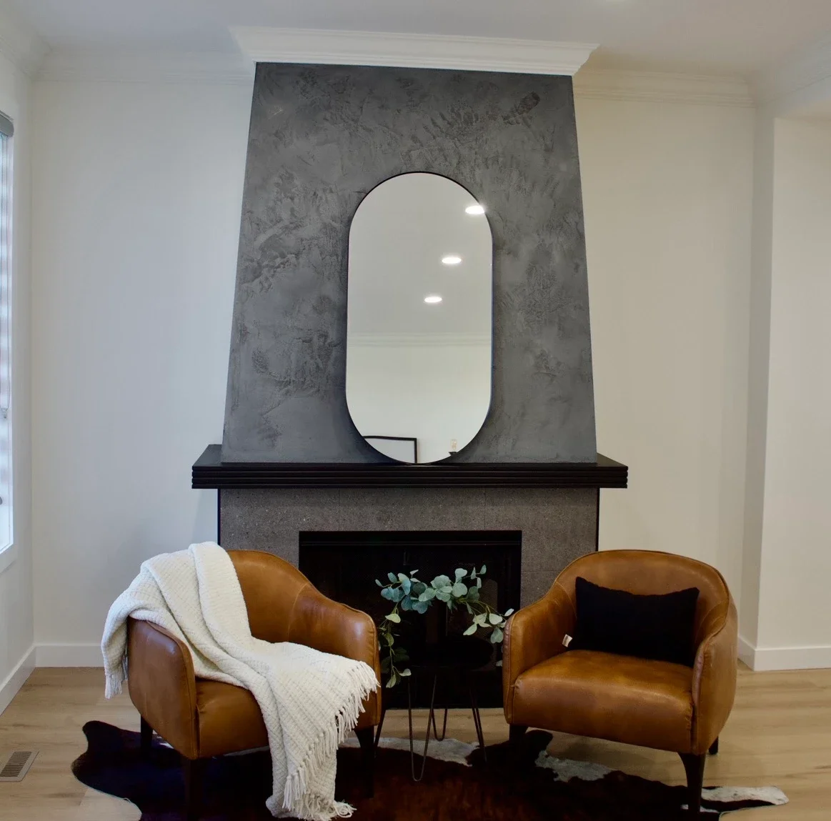 Living room with a fireplace, a large mirror above the mantle, two brown leather armchairs, a small black table with greenery, a white blanket on one chair, a black pillow on the other, and a cowhide rug on the wooden floor.
