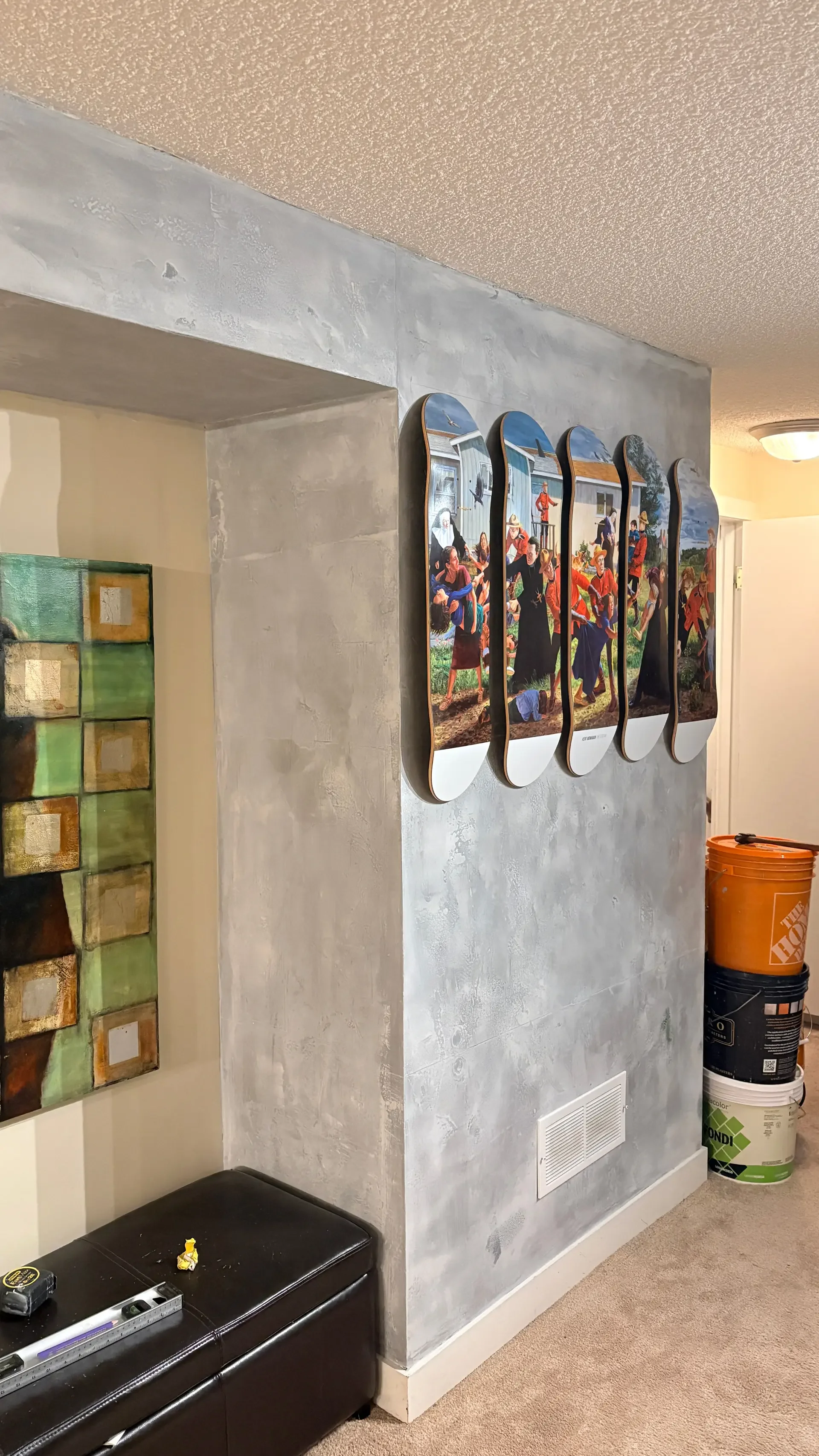 A set of five skateboards mounted on a textured gray wall inside a home, each featuring a painting of a group of people in a rural setting.