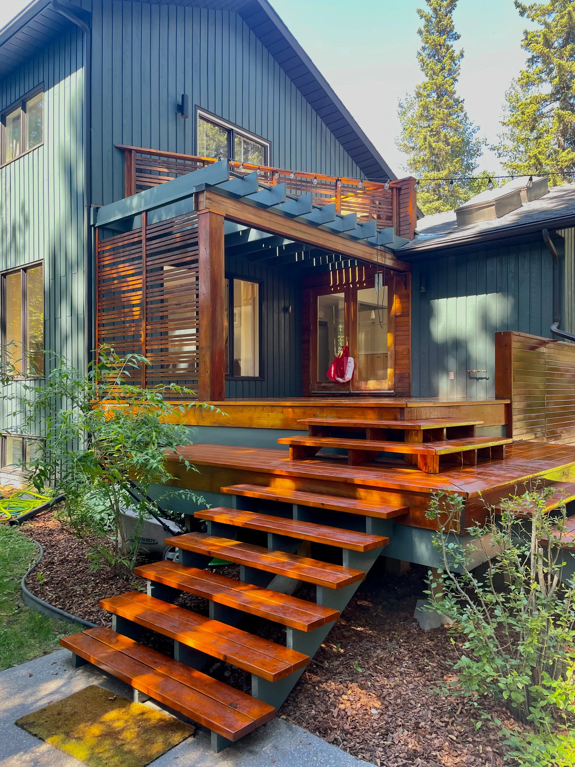 Back porch of a modern house with wooden stairs leading up to the door, featuring blue siding and a small balcony with wooden railing, surrounded by trees and greenery.