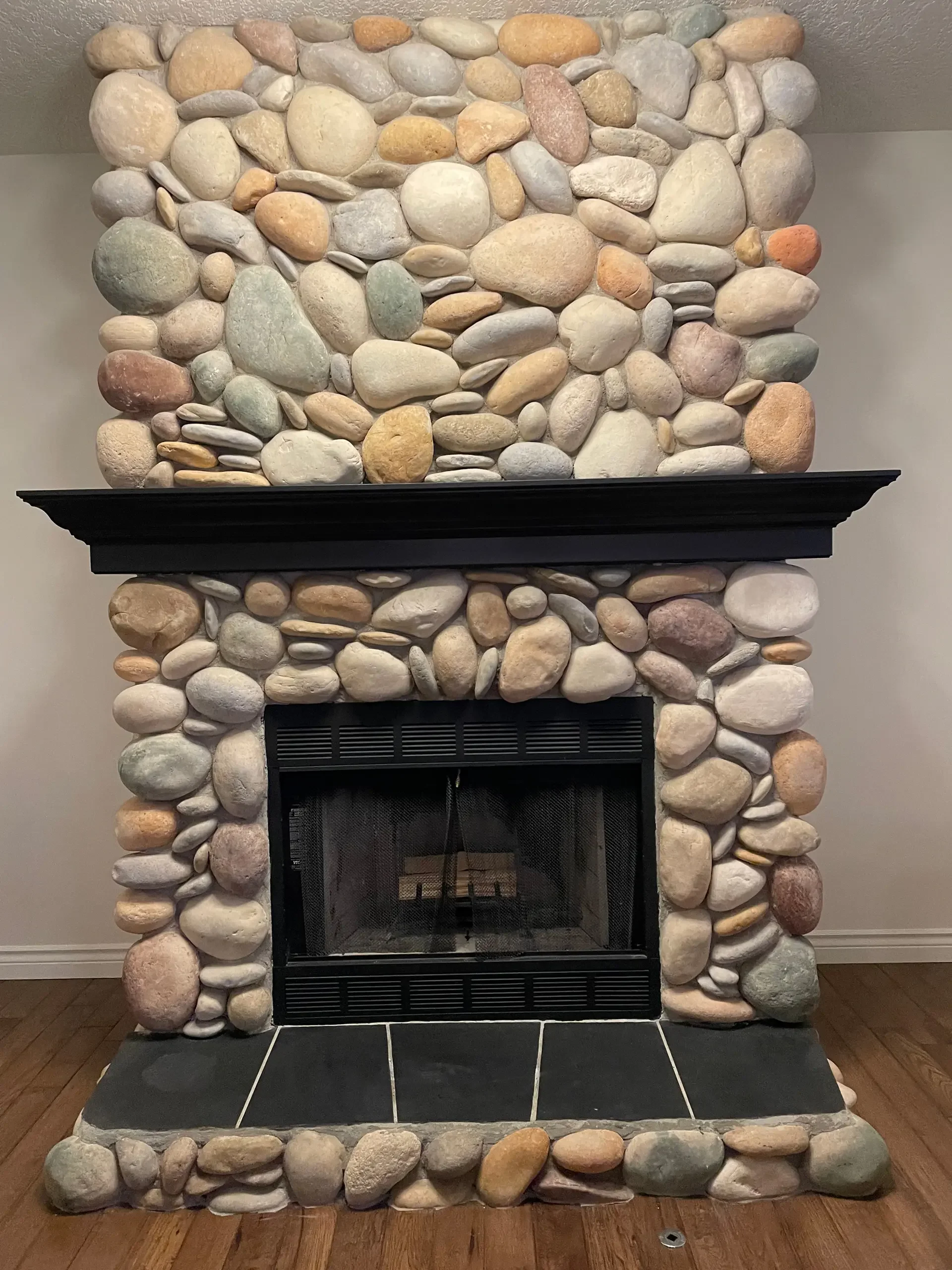 A stone fireplace made of rounded river rocks with a black mantel and a black tiled hearth. The rocks vary in color from white, beige, gray, brown, and some with hints of pink.