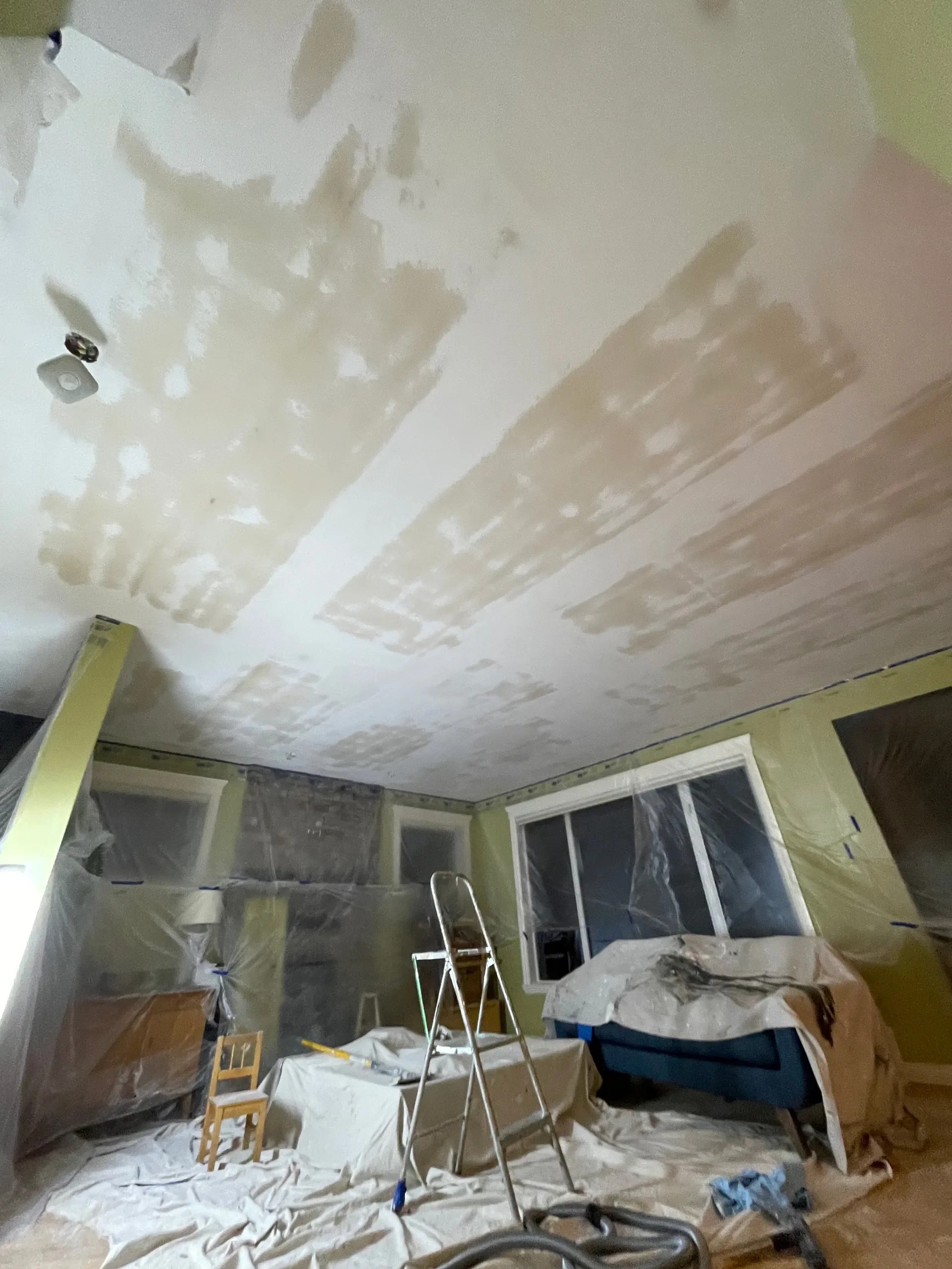Room undergoing painting or drywall work with plastic covering furniture and walls, ladder in the center, and a table covered with a cloth.
