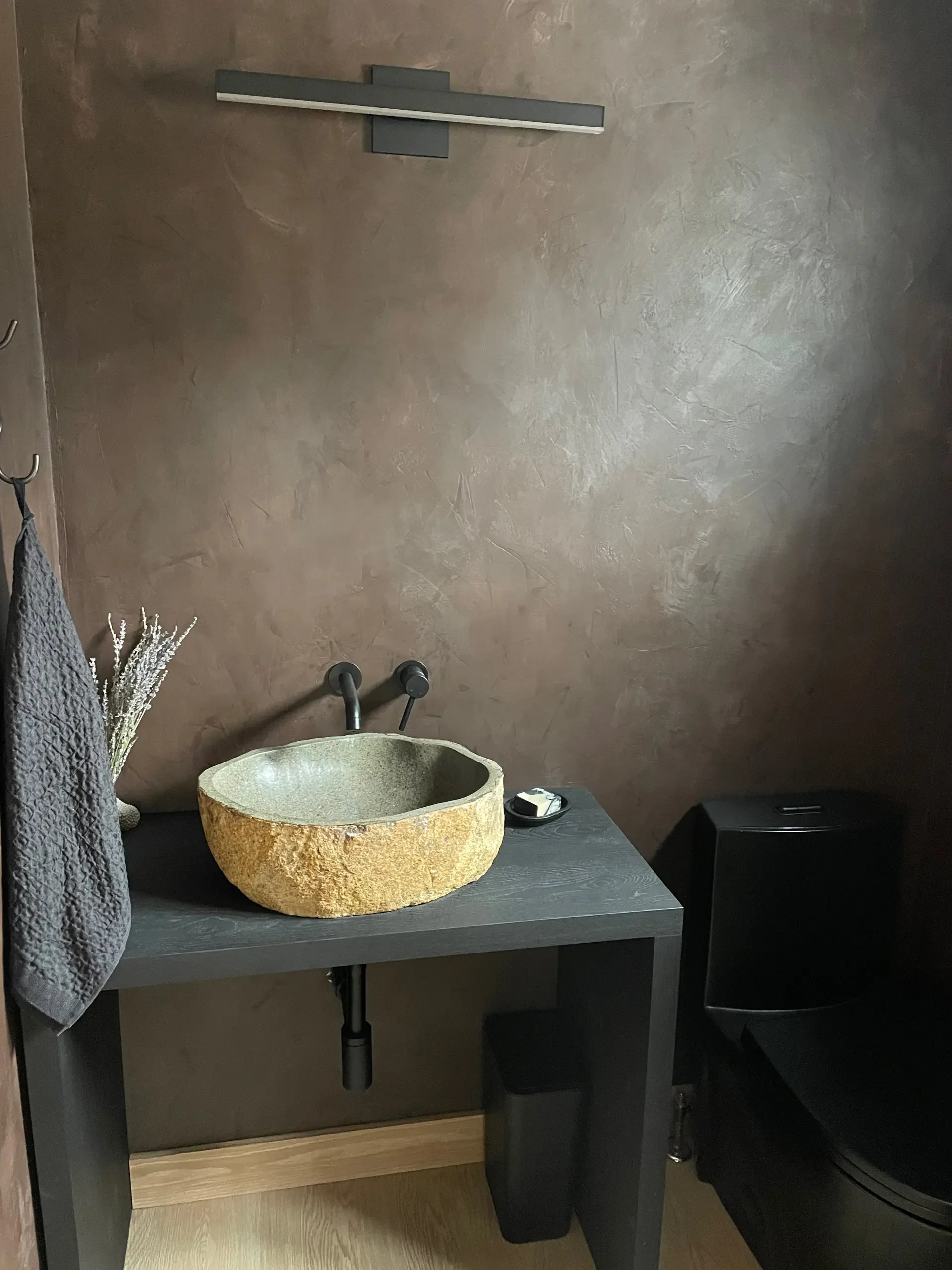 Bathroom with a stone sink on a black table, a black toilet, and a beige wall with a modern silver light fixture.