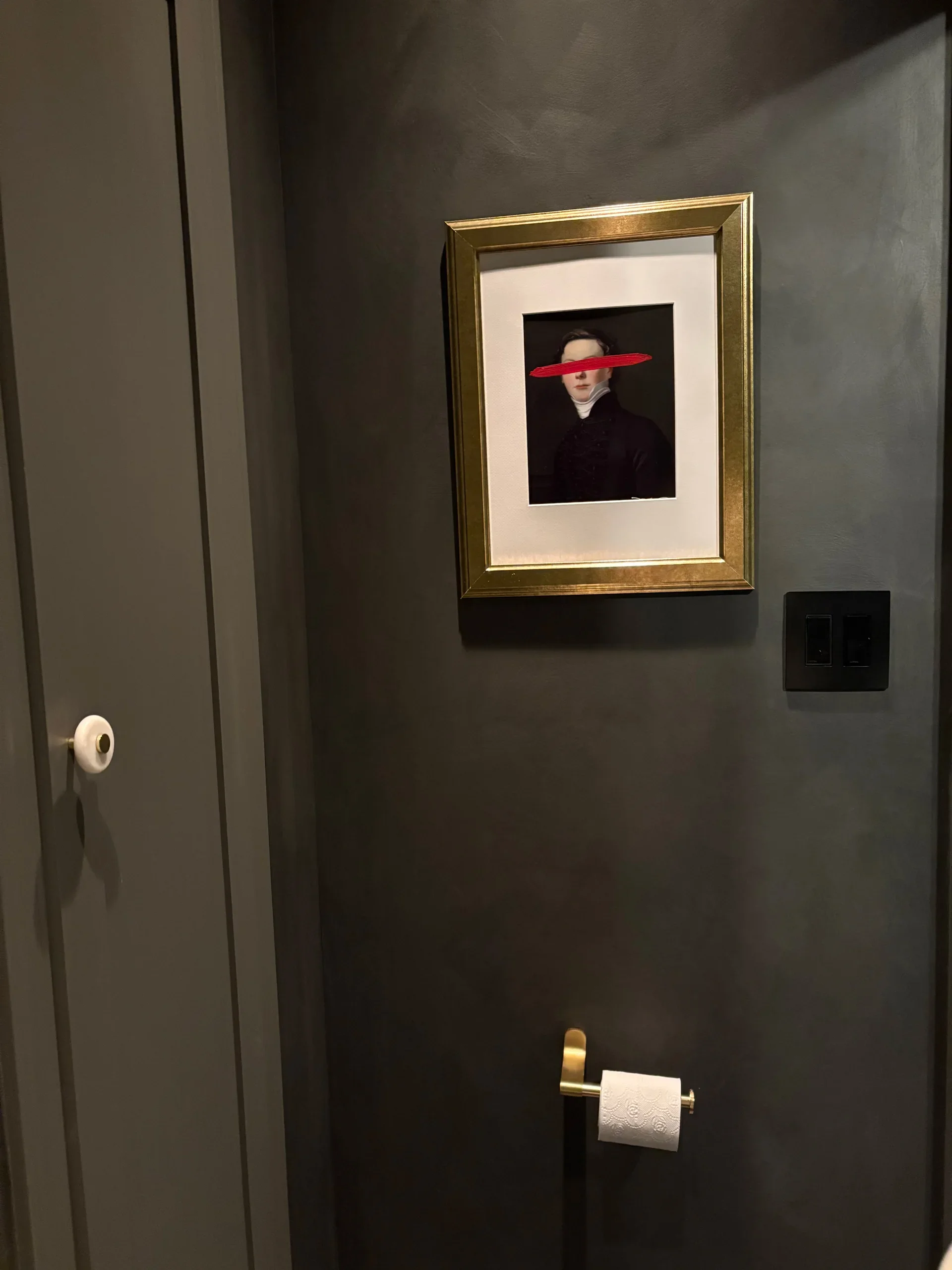 A framed portrait of a woman with dark hair styled in a vintage fashion, dressed in black with a white high-neck collar, has a red brushstroke obscuring her eyes, hanging on a dark gray wall in a room with a black light switch and a gold toilet paper