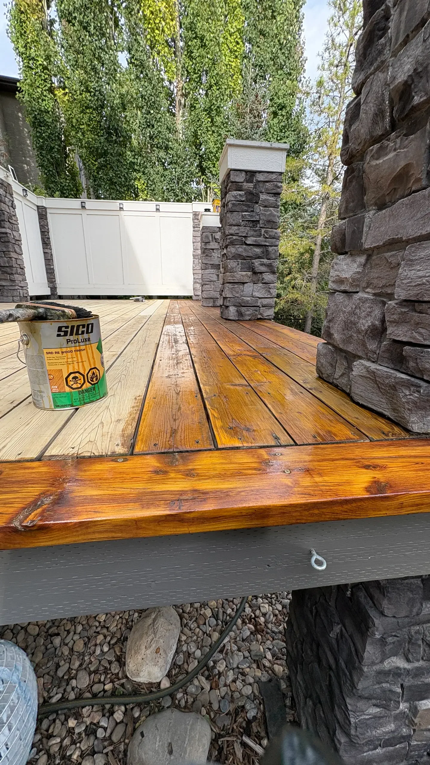 Close-up of a wooden deck under construction, with a can of wood stain or finish, stone pillars, a white fence in the background, and trees surrounding the area.