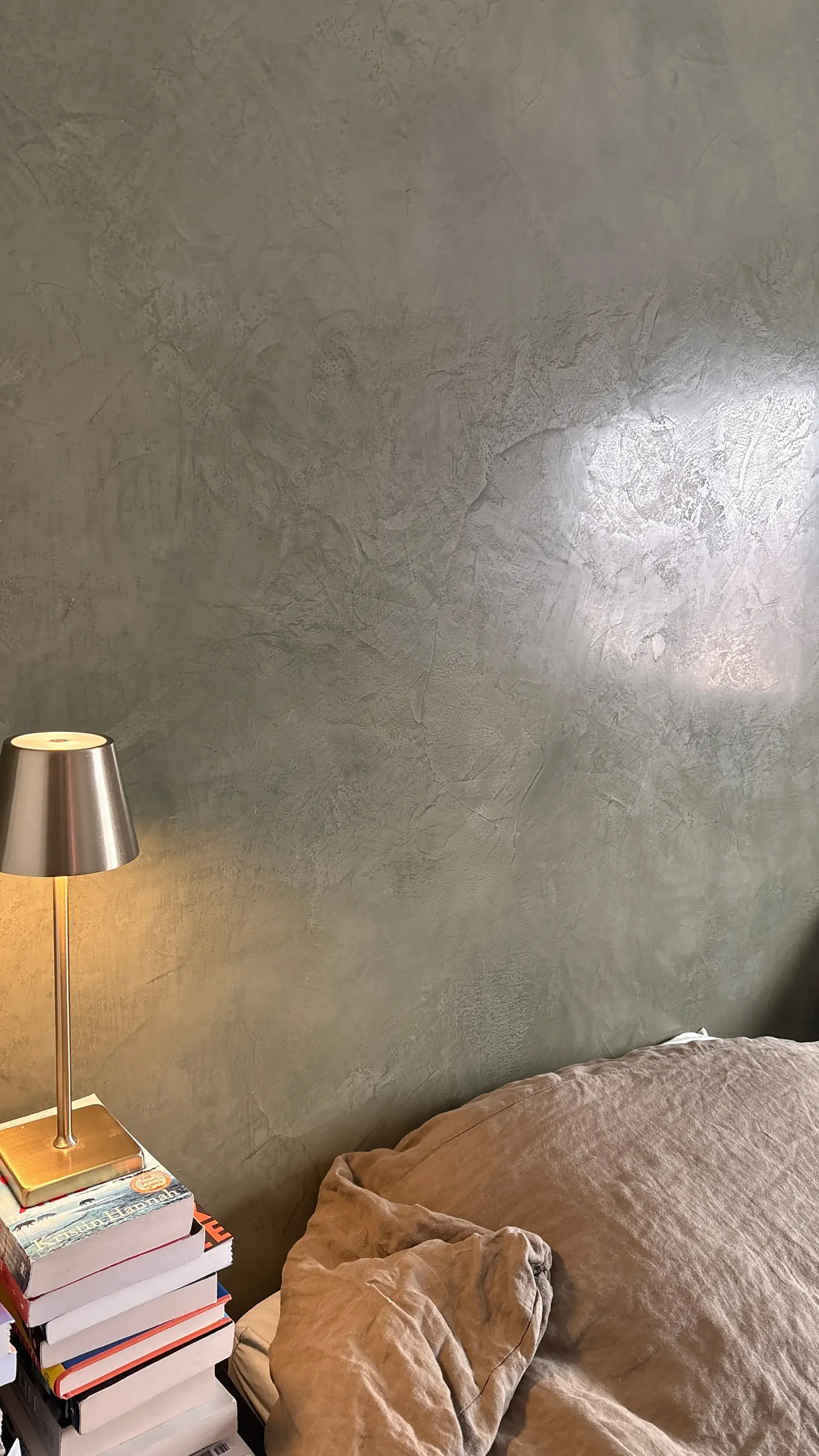 A bedroom scene showing a textured gray wall, a bed with beige bedding, a bedside lamp, and a stack of books.